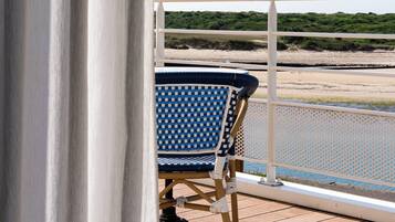 Un balcón con una silla azul y blanca a cuadros, barandilla blanca y vista a la playa y las dunas.
