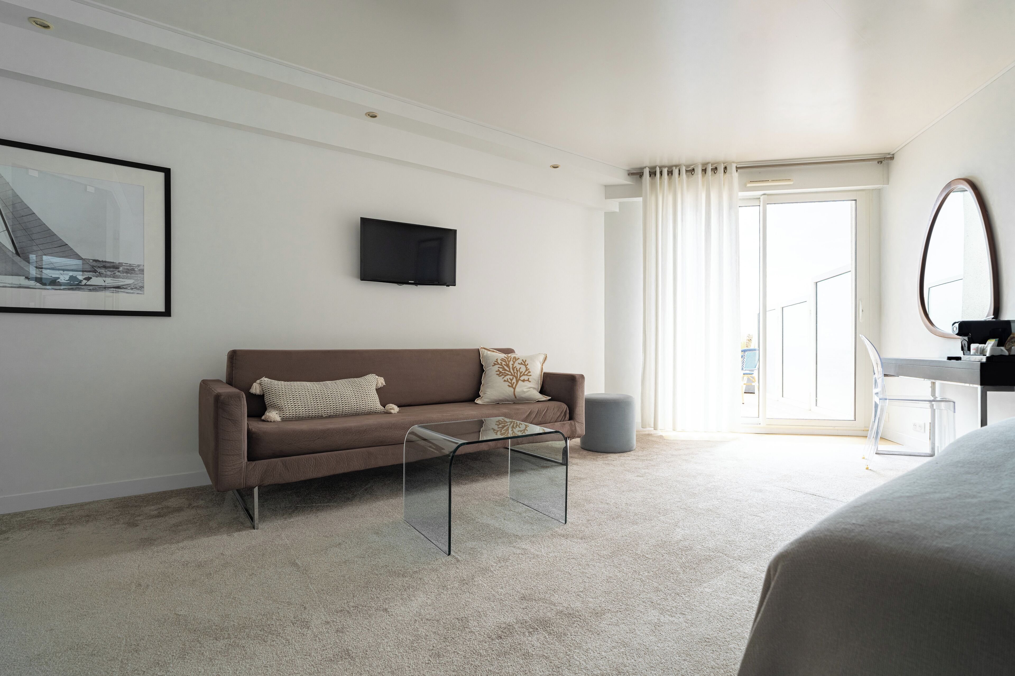 Habitación de hotel moderna con un sofá marrón, una mesa de centro de vidrio, un televisor de pantalla plana, un espejo grande y vistas al exterior a través de una ventana con cortinas.