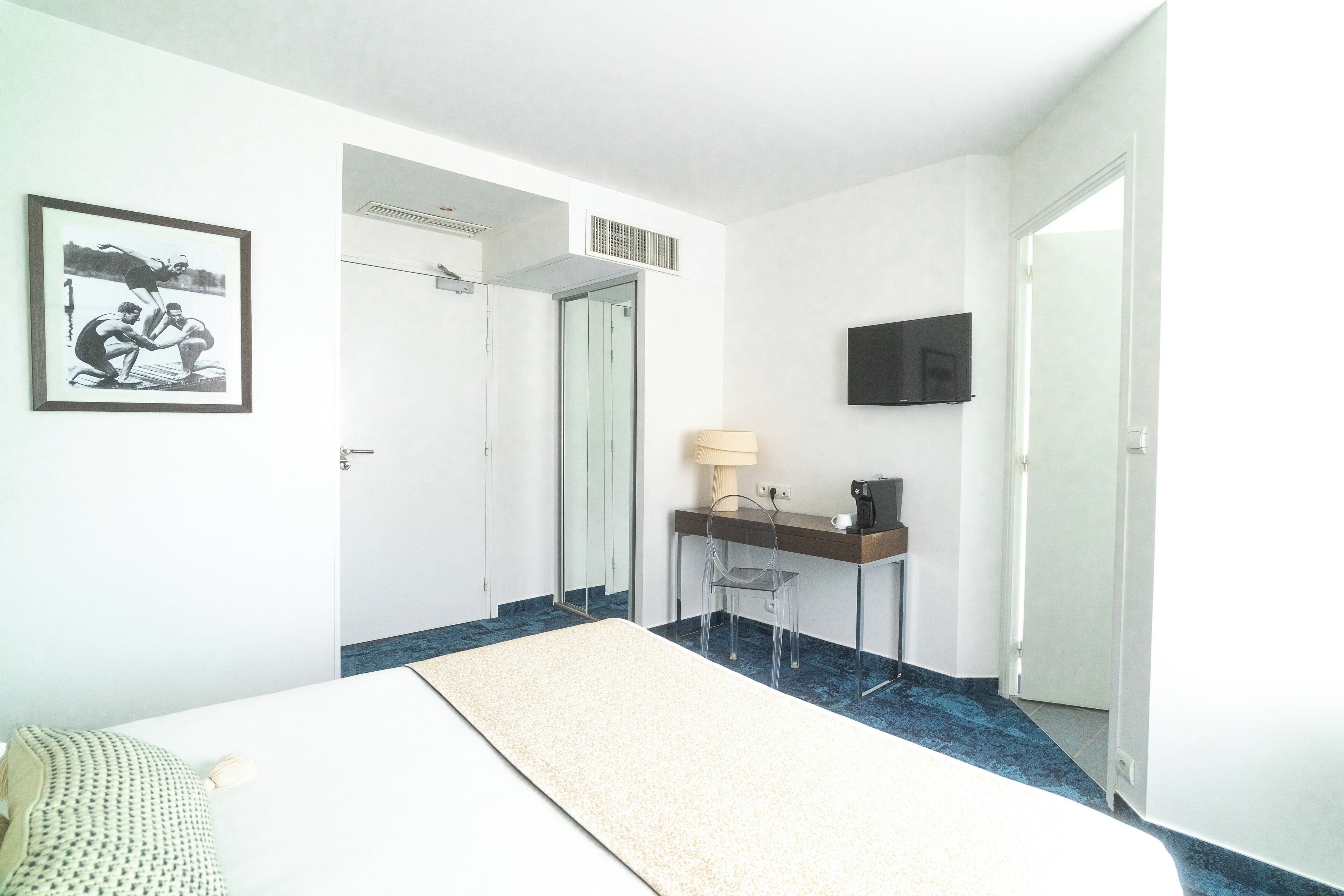 A hotel room with a bed, a desk with a lamp, a TV, and a framed picture on the wall.