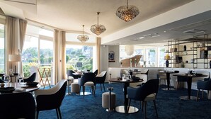 A modern dining area with round tables and chairs, large windows with curtains, and decorative hanging lights. - La Marine (Barneville-Carteret)