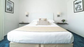 Una cama bien hecha con colcha blanca y beige, dos mesitas de noche con lámparas, y cuadros en las paredes.