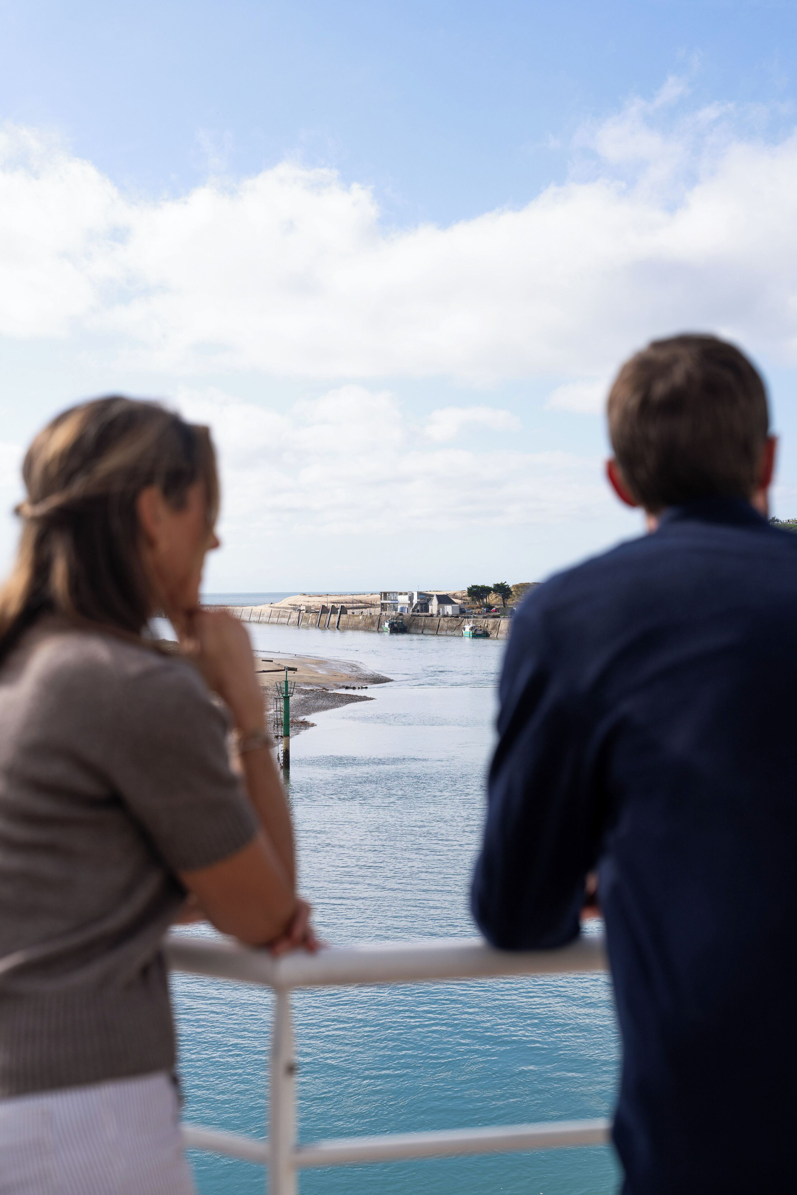 Two people overlooking a waterfront with a bridge and industrial structures in the background.