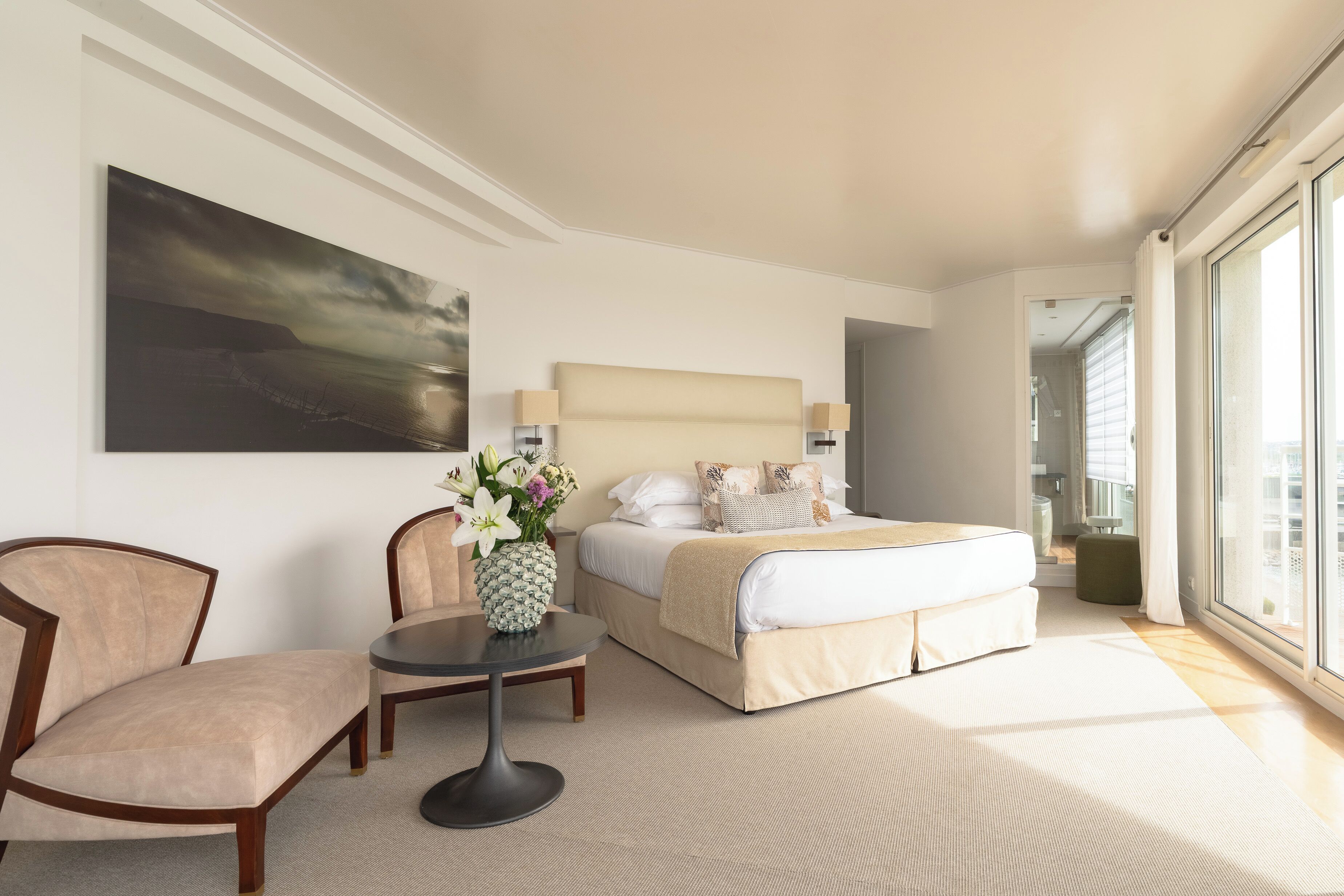 Une chambre d’hôtel avec un lit, une chaise, une petite table sur laquelle se trouve un vase, et une grande fenêtre.