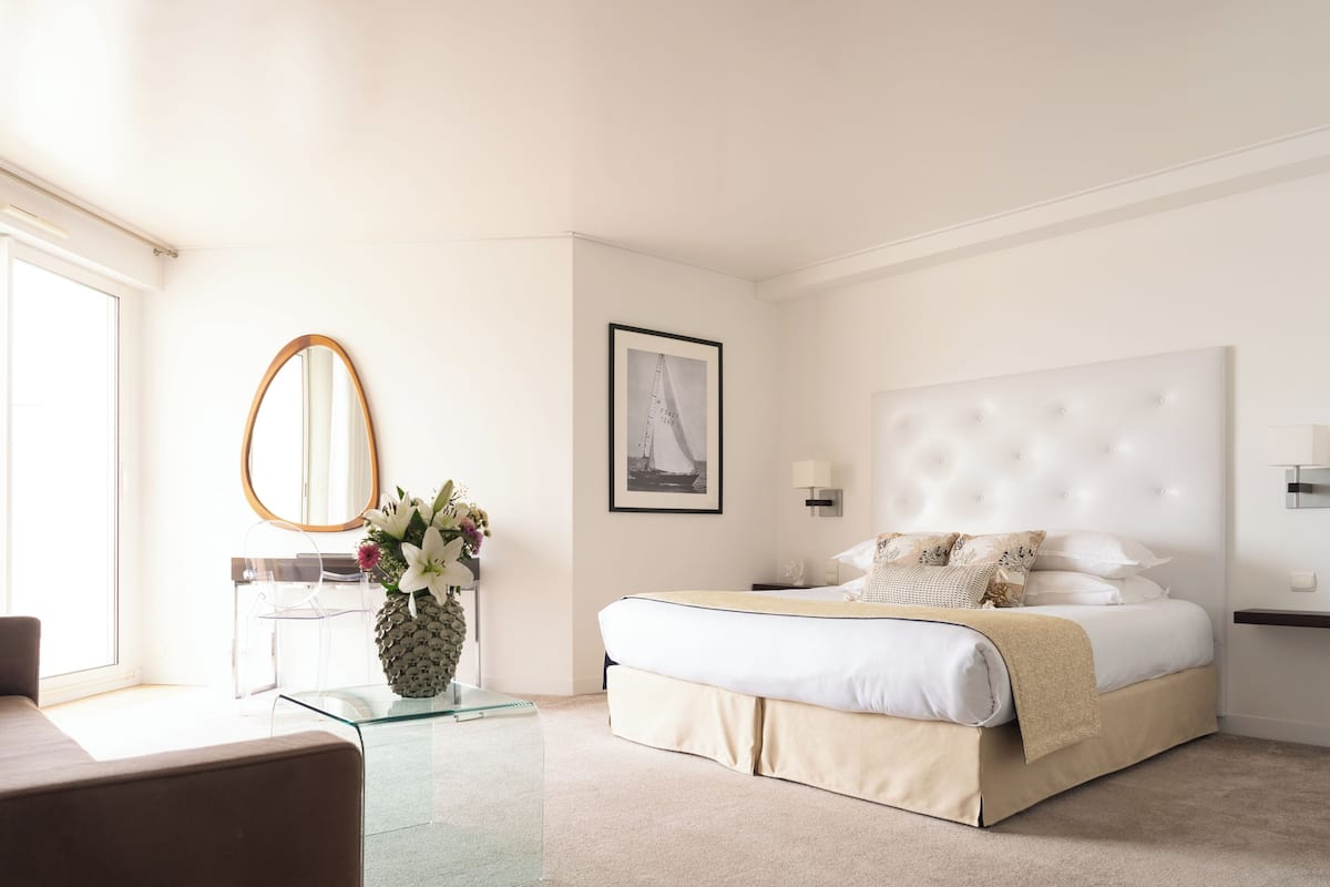 A hotel room with a bed, a bedside table, a mirror, a framed picture on the wall, and a window.