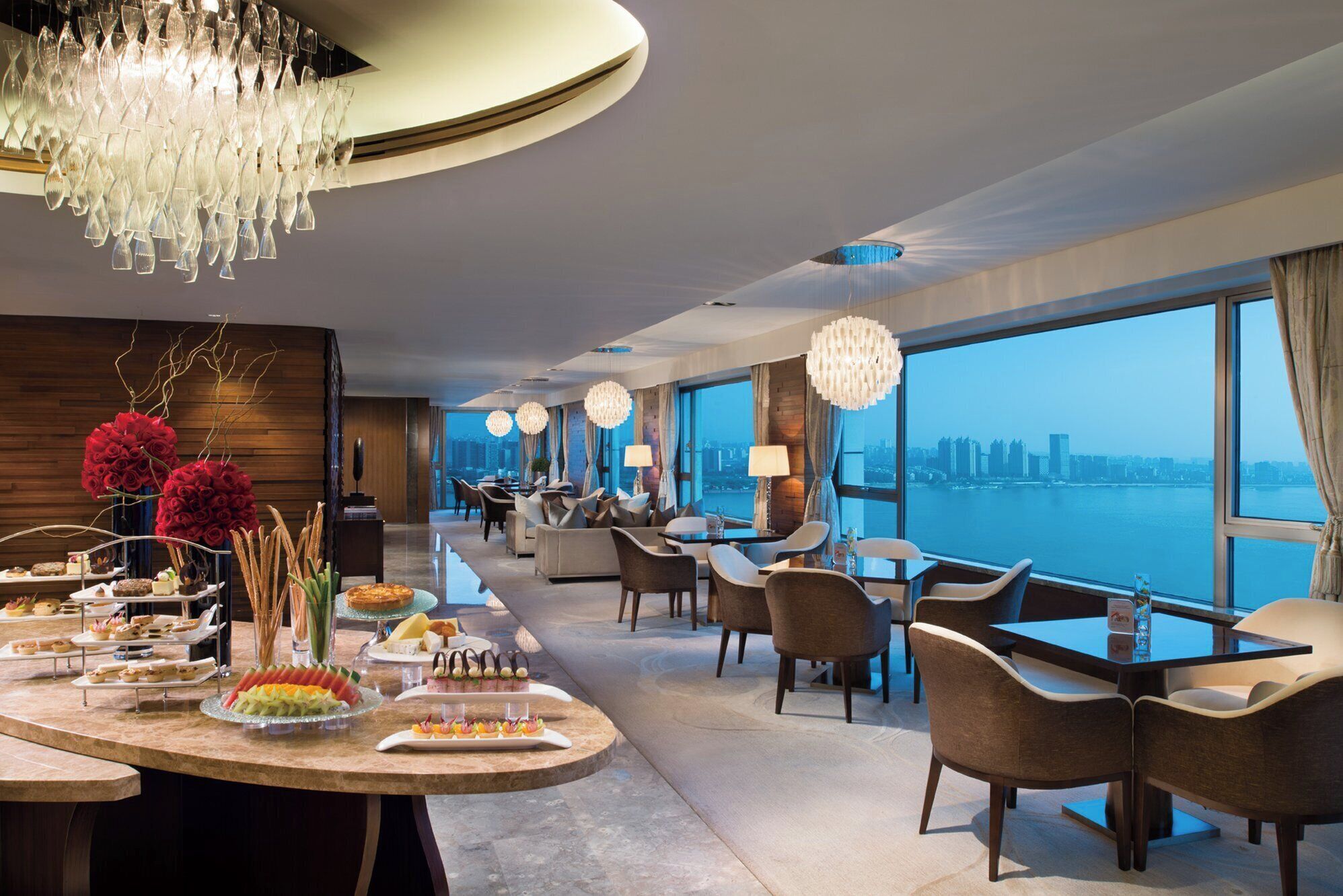 A hotel dining area with a long counter displaying various desserts and a view of the cityscape through large windows.