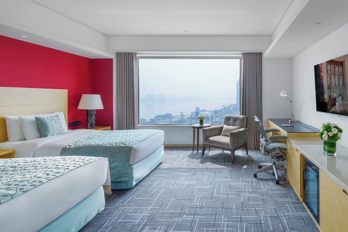 Een hotelkamer met twee bedden, een bureau, een stoel en een groot raam met uitzicht op de stad.