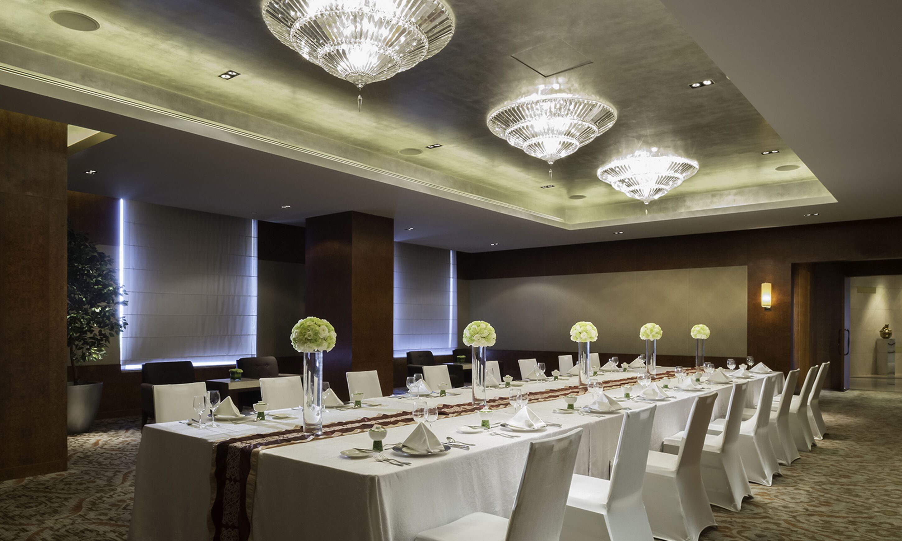 Une salle de banquet avec de longues tables dressées pour le repas, des lustres et un design de plafond moderne.