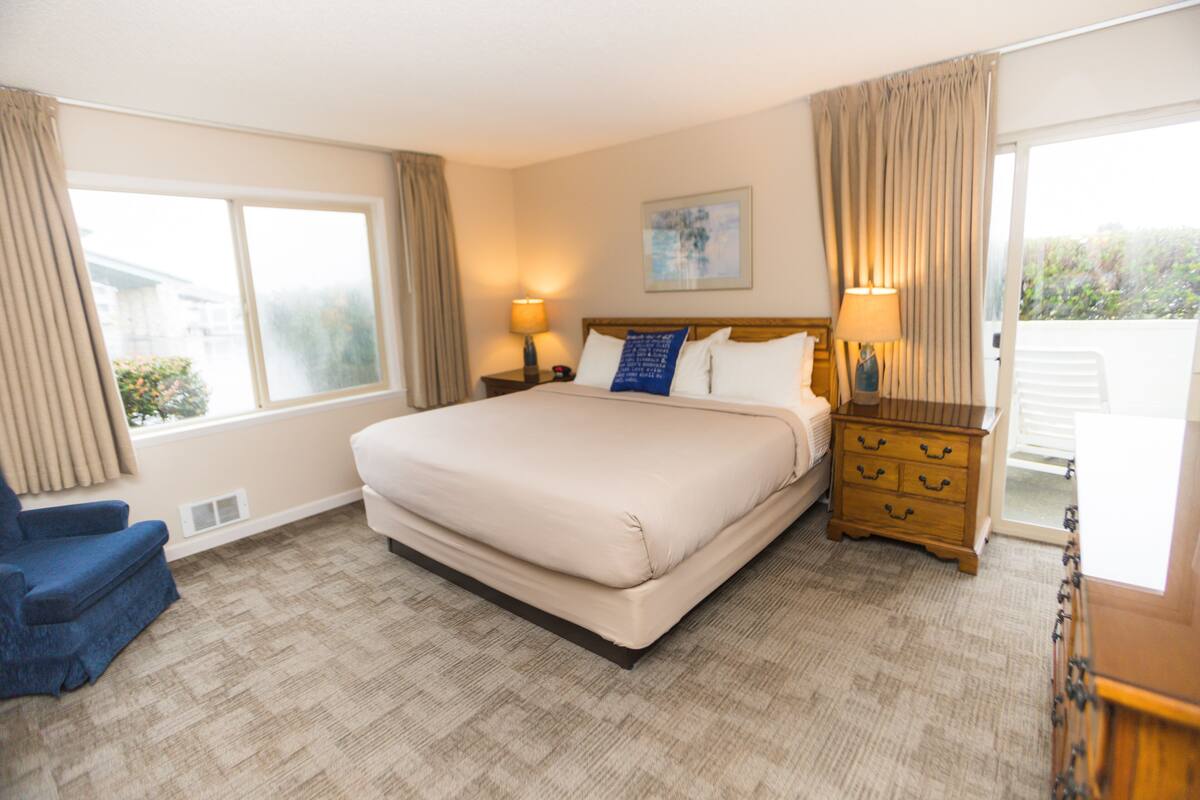 Habitación de hotel con una cama grande, una silla, un aparador y un ventanal con cortinas.