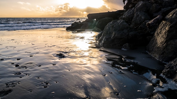 A beach with rocks and waves, and the sun is setting in the background.