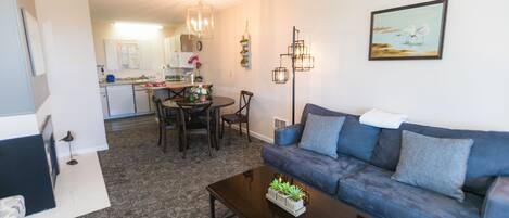 A modern living room with a blue sofa, a coffee table with plants, a dining area with a table and chairs, and a kitchen in the background.