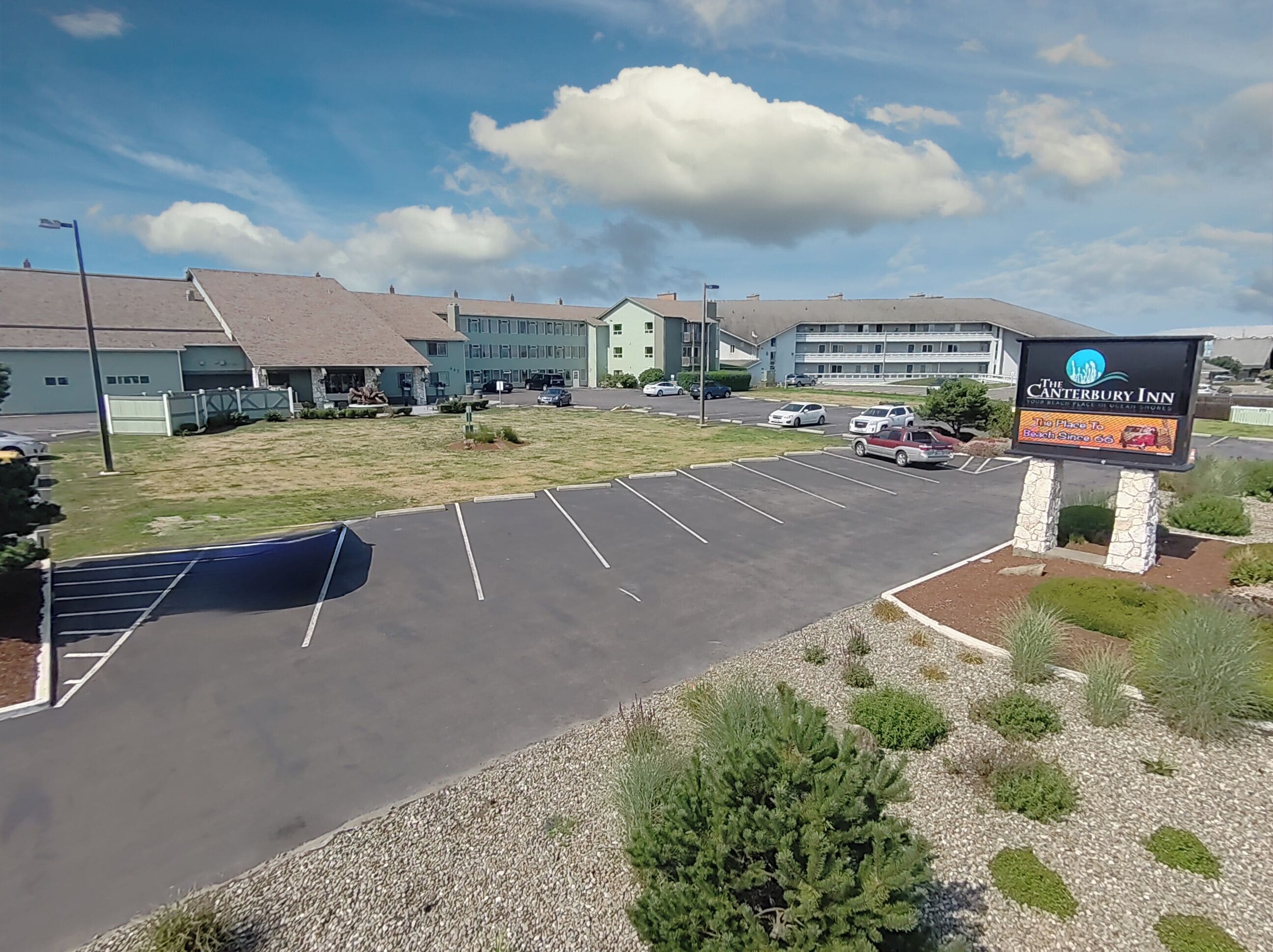 A parking lot with a sign for The Canterbury Inn, a building with a flat roof, and a grassy area.