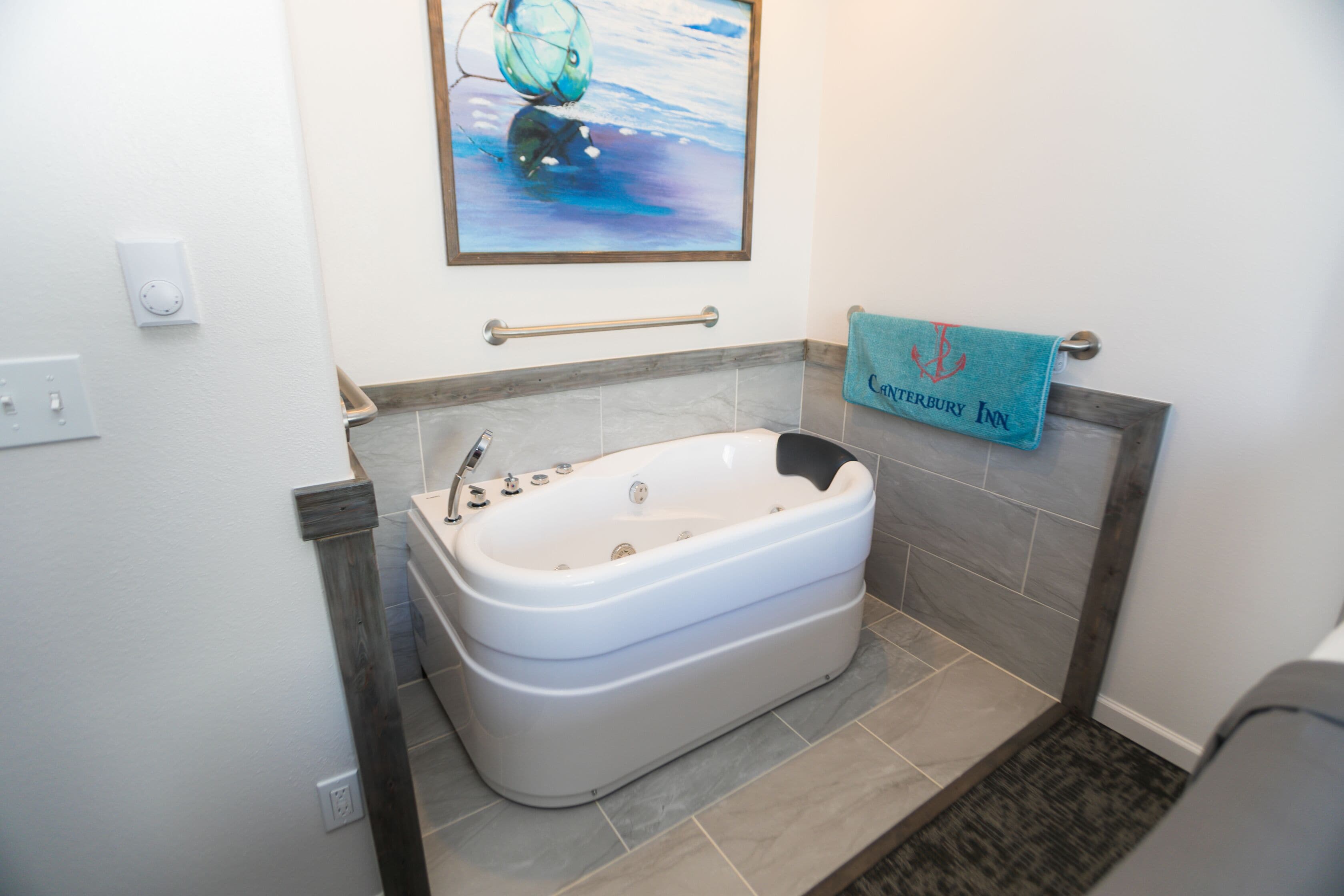 Ein Badezimmer mit einer großen Badewanne, einem Handtuchhalter mit Handtuch, einem Bild an der Wand und einer Haltestange.