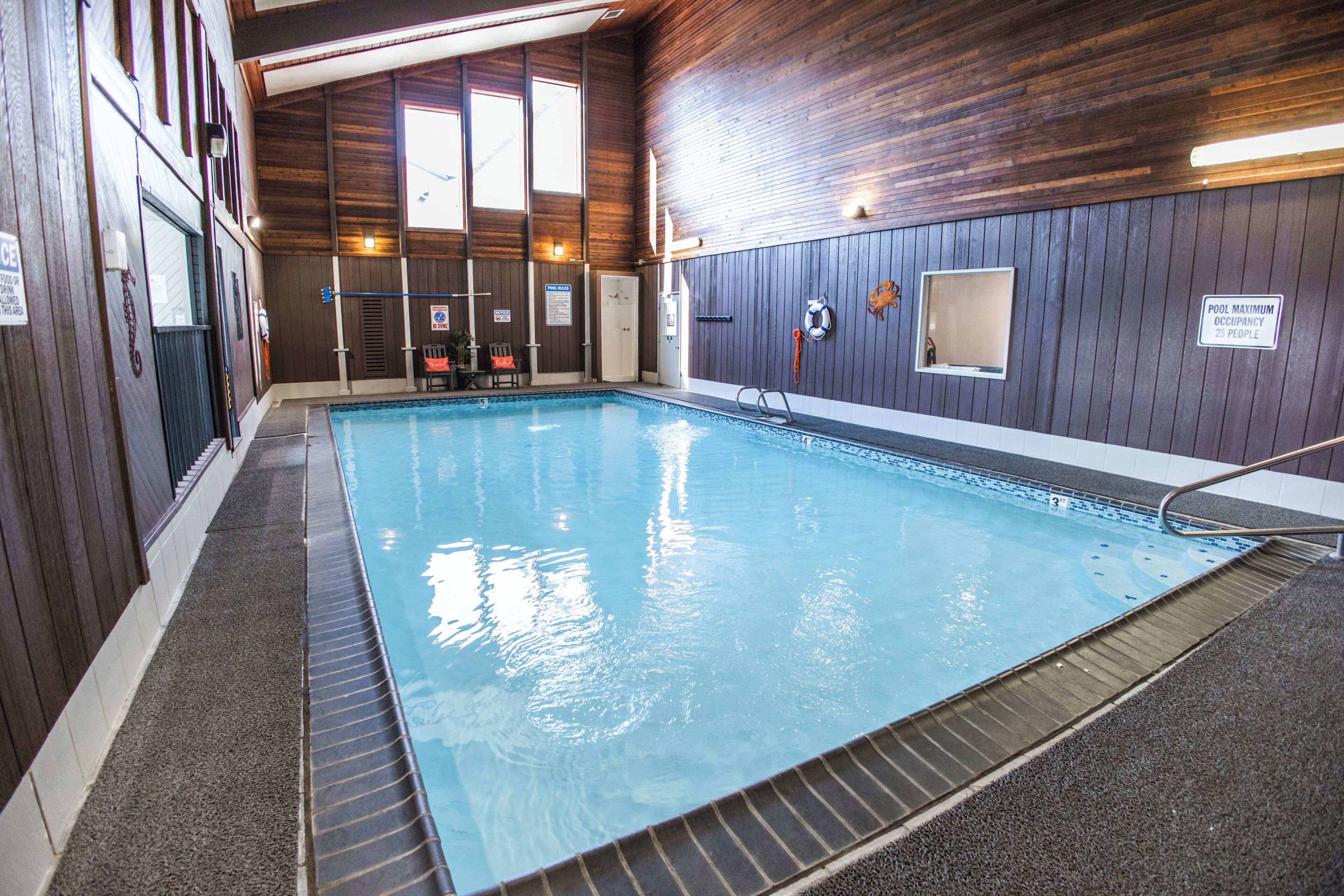 Une piscine intérieure aux murs en bois, dotée d’un poste de surveillance pour les sauveteurs et d’un panneau indiquant une capacité maximale de 25 personnes.