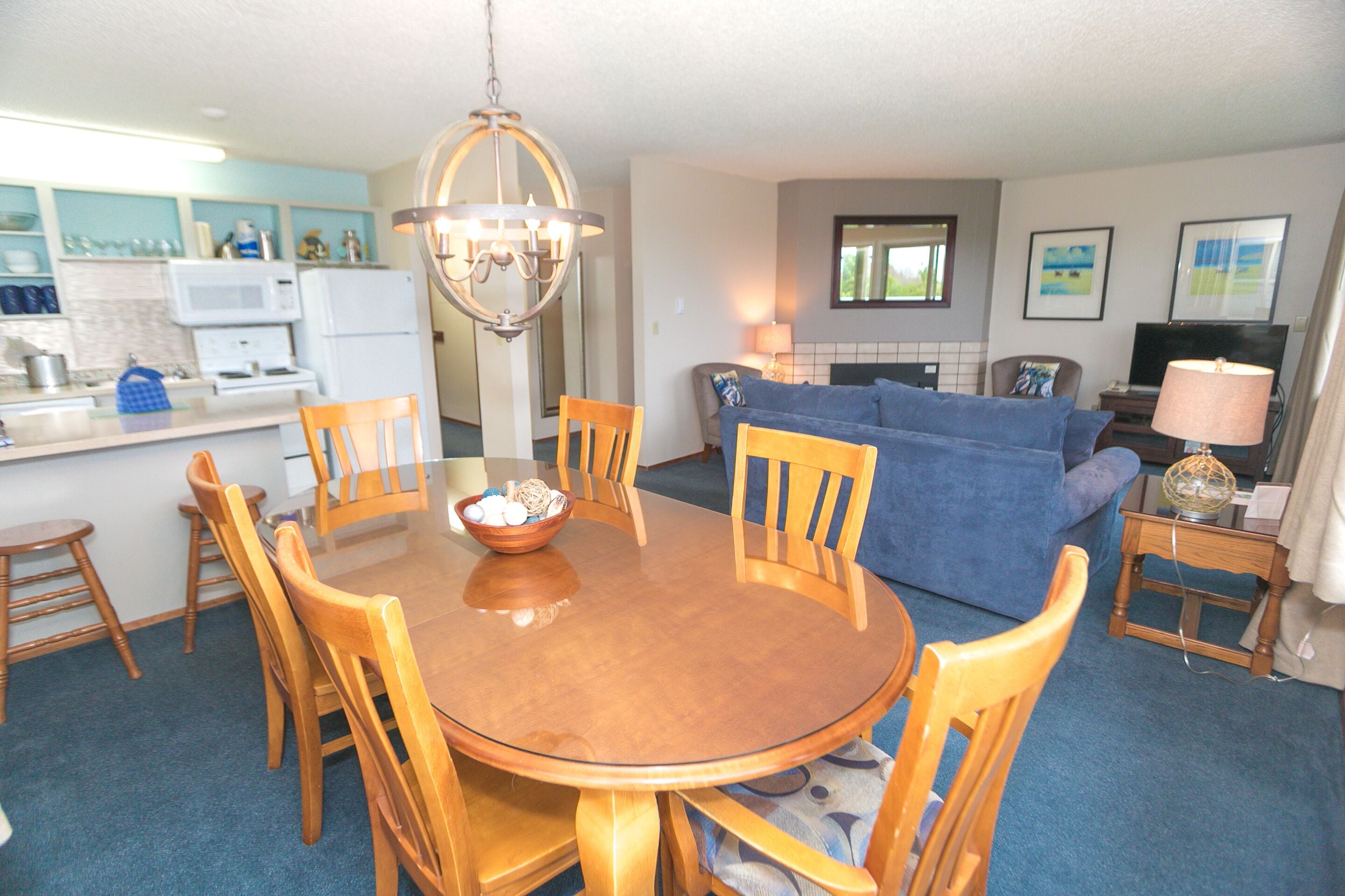 A dining area with a round wooden table, wooden chairs, and a chandelier. There is a kitchen area with a microwave and a refrigerator in the background.