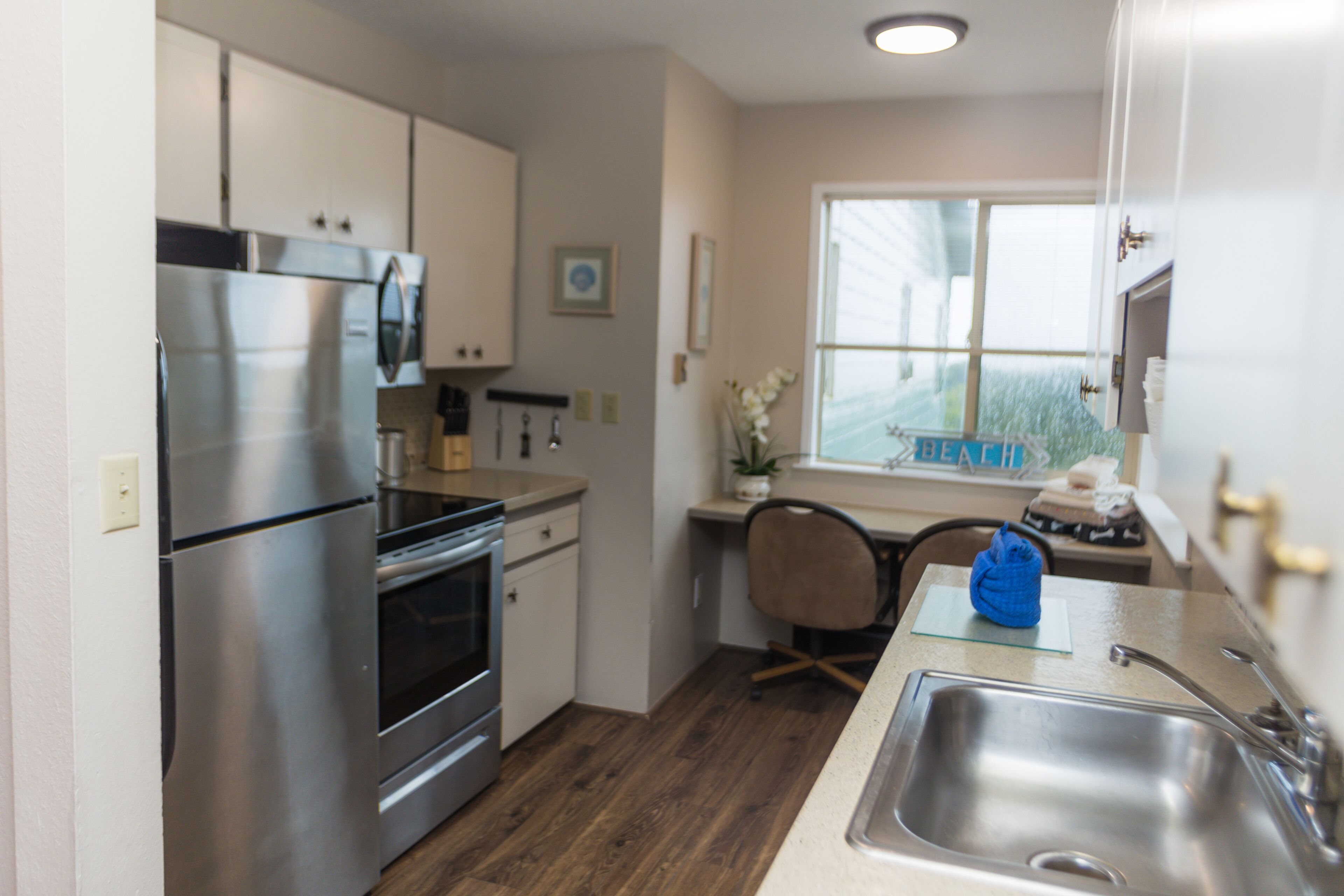 A modern kitchen with stainless steel appliances, a wooden floor, and a window with a view of the beach.
