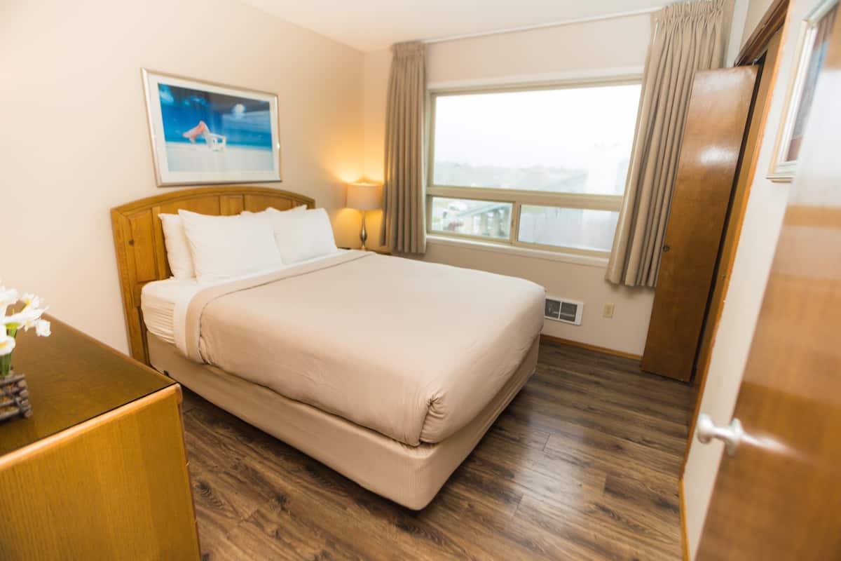 Habitación de hotel con una cama grande, cabecera de madera, una mesita de noche con lámpara, ventana con cortinas y un cuadro en la pared.
