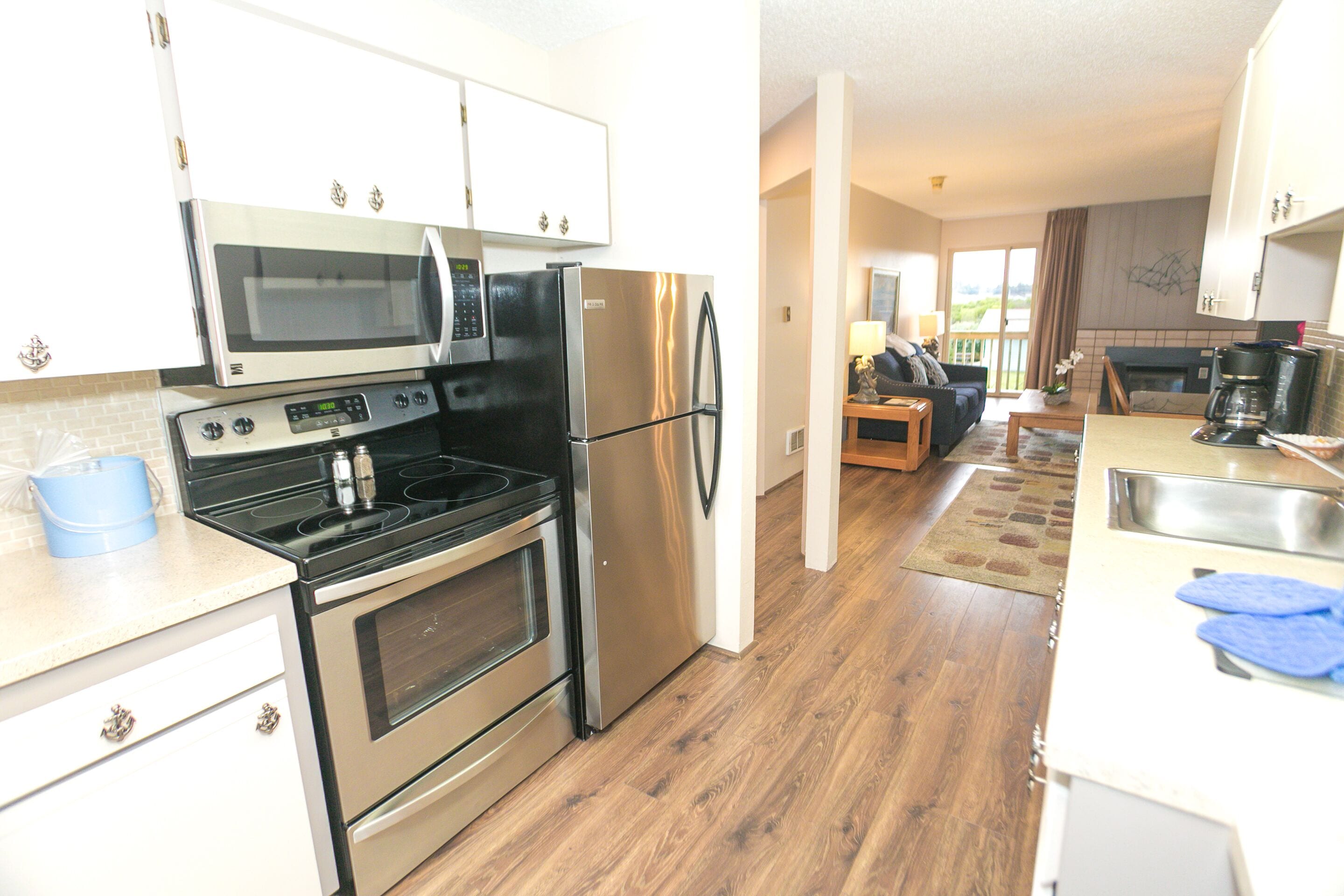 A modern kitchen with stainless steel appliances, wooden flooring, and a view to the living area.