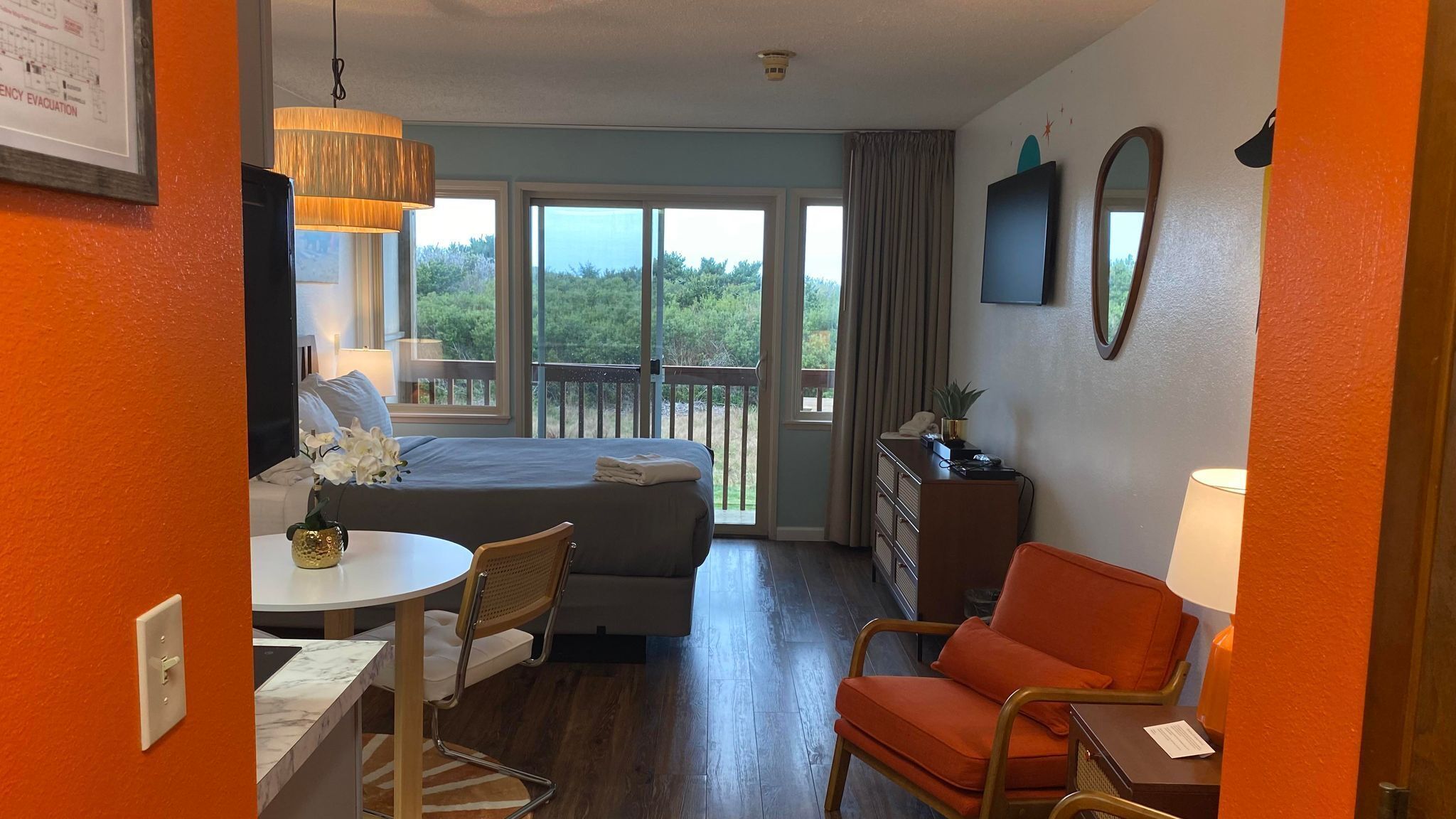 A hotel room with a bed, a desk with a chair, a dresser, and a balcony with a view of greenery.