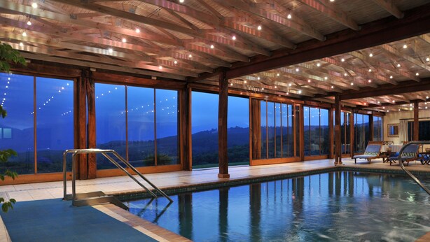An indoor swimming pool with a view of mountains, wooden beams, and a poolside area with chairs. - Coach House Hotel & Spa (Tzaneen)