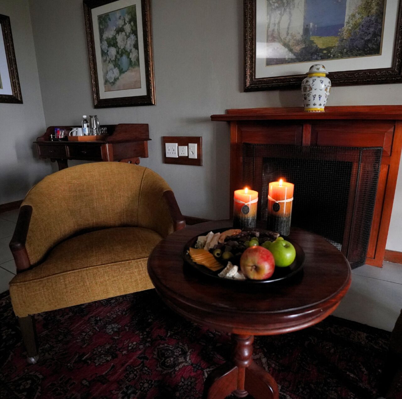 A cozy living room with a wooden armchair, a small round table with fruit and candles, and a fireplace with a vase on top.