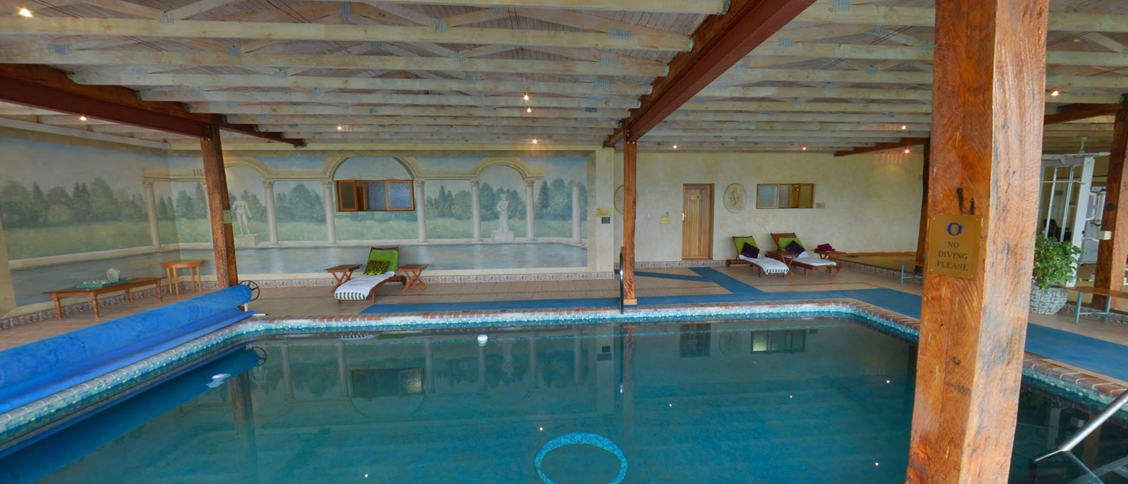 Una piscina interior con un mural, sillas de descanso y una estructura de techo de madera.