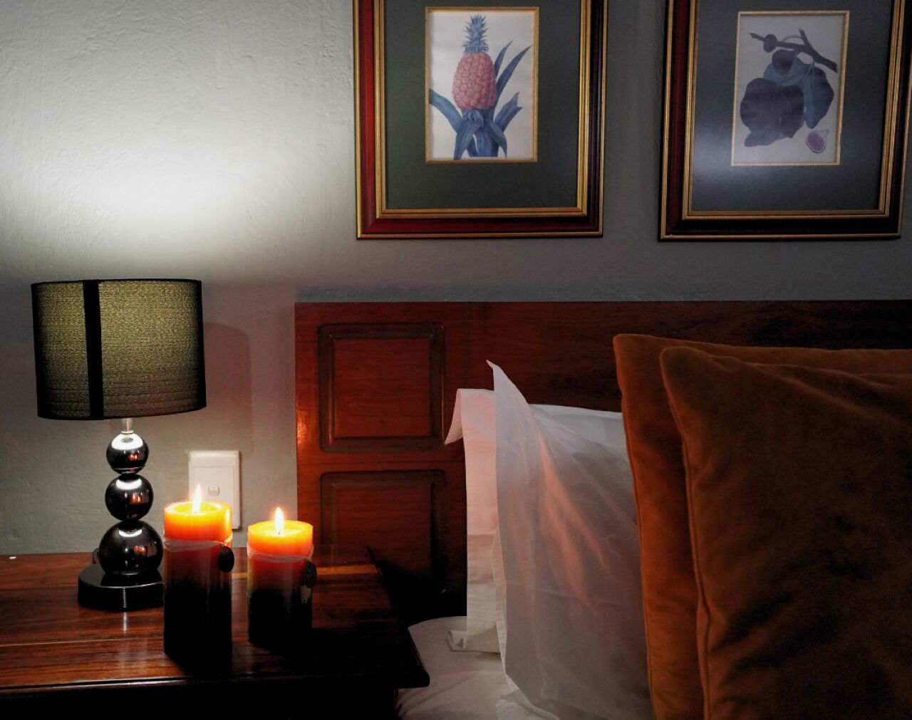 Habitación de hotel con cabecera de madera, mesita de noche con lámpara, dos velas encendidas y una obra de arte enmarcada en la pared.