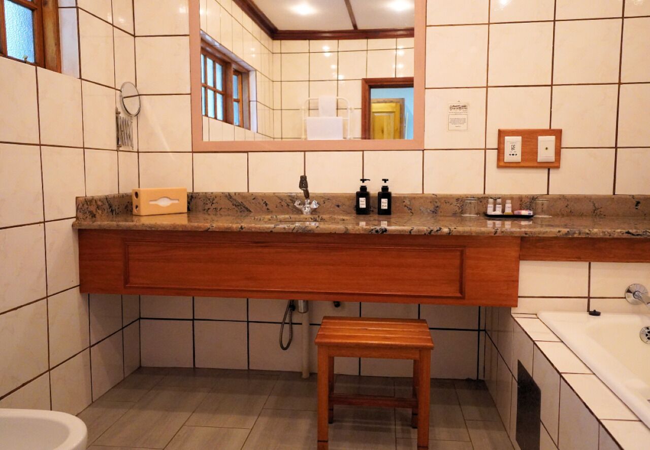 Un baño con encimera de mármol, tocador de madera, un lavabo con grifo, un espejo, un taburete de madera pequeño, una bañera y paredes con azulejos.