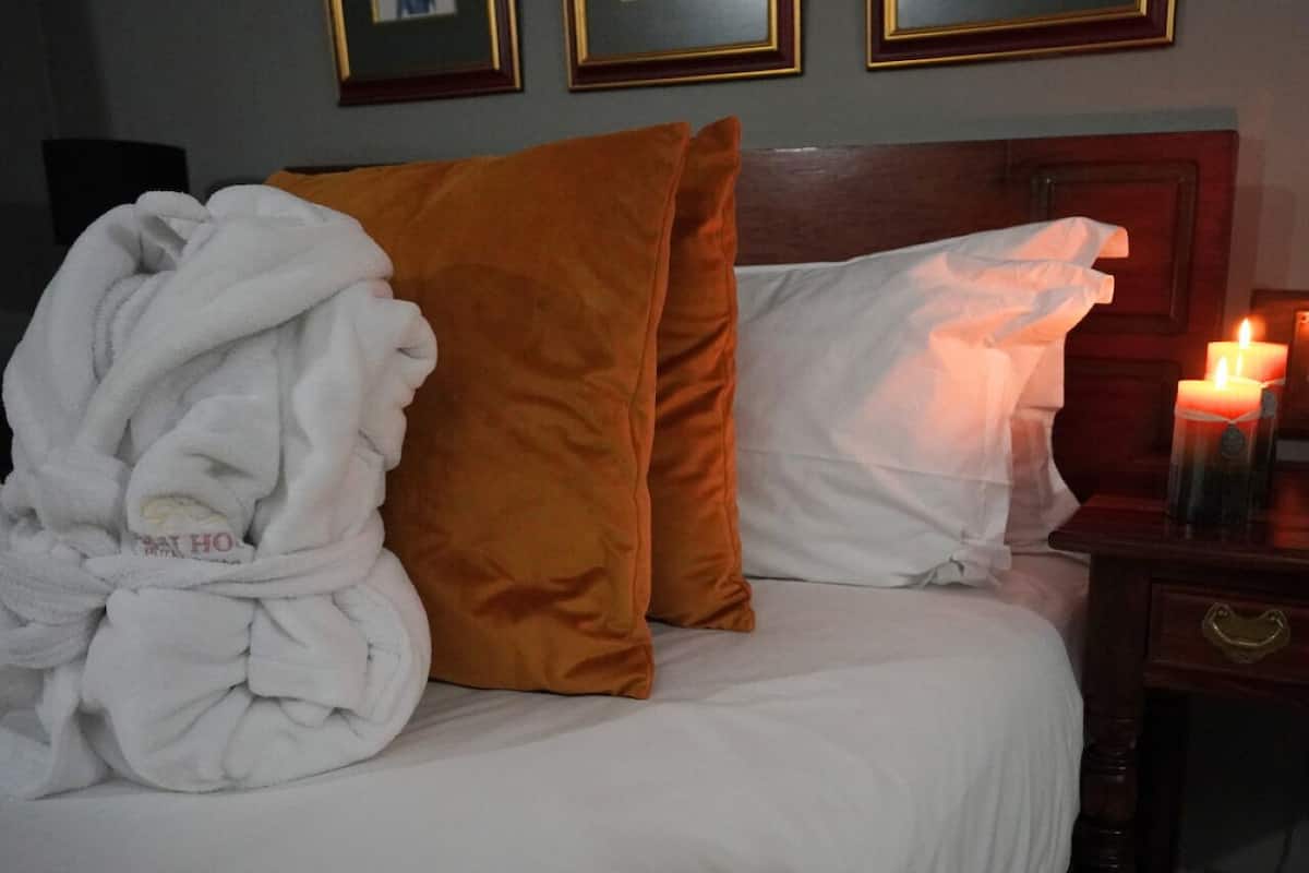 Ein ordentlich bezogenes Bett mit weißen und orangefarbenen Kissen, einem weißen Bademantel und einer Kerze auf einem Nachttisch.