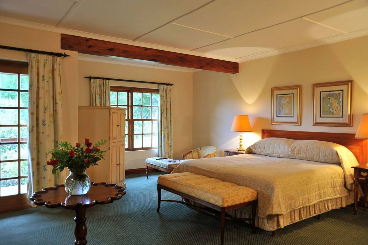 Ein Hotelzimmer mit einem großen Bett, einer hölzernen Bank, einem Schreibtisch, einem Stuhl, einem Schrank und einer Blumenvase.