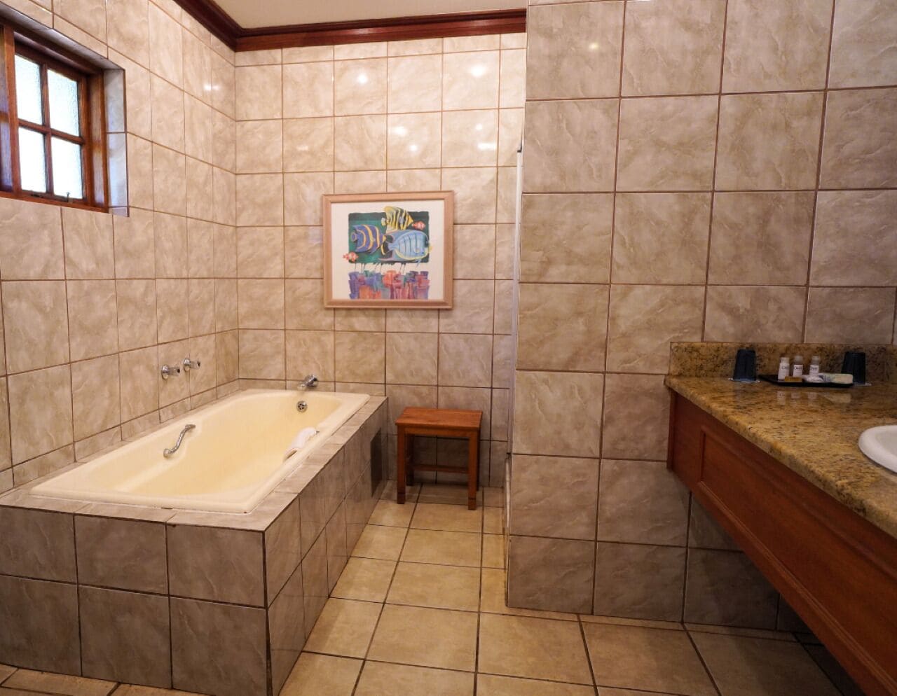 Un baño con una bañera grande, un taburete de madera, una obra de arte enmarcada en la pared y una encimera de granito con un lavabo.