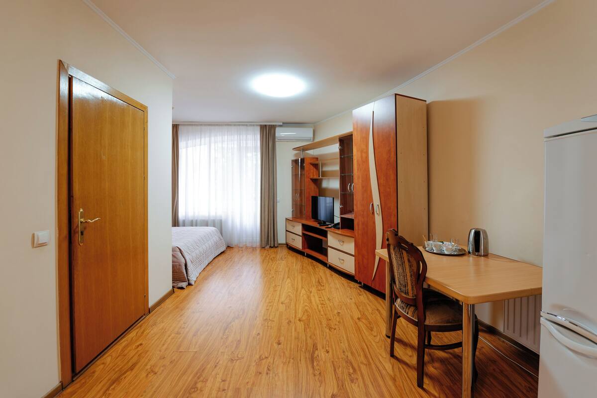 A compact hotel room with a wooden floor, a bed, a wardrobe, a desk with a chair, and a refrigerator.
