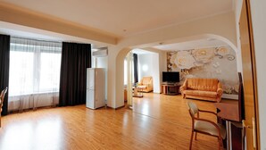 Een ruime woonkamer met houten vloeren, een witte koelkast, een bank, een televisie en een eettafel met stoelen.