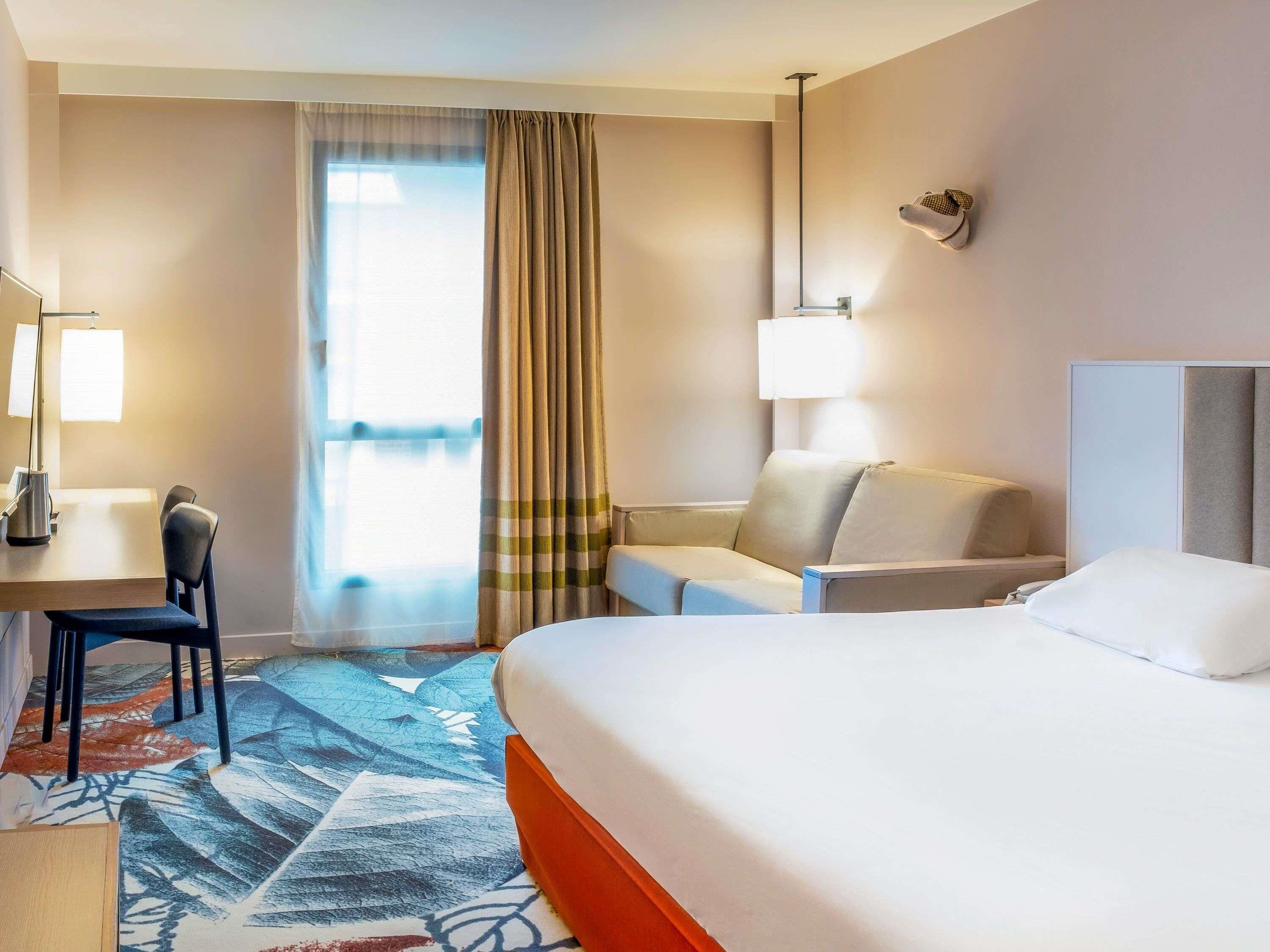A hotel room with a bed, a desk with a chair, a sofa, and a large window with curtains.