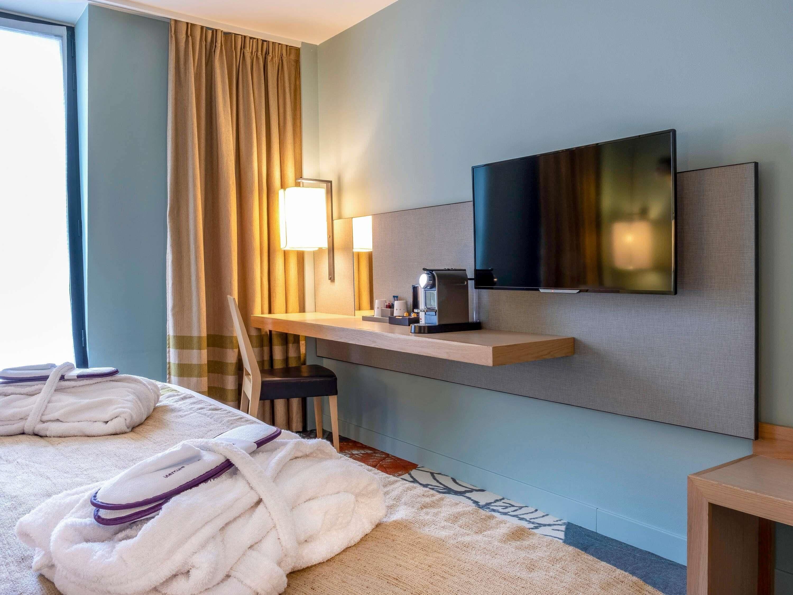 A hotel room with a bed, a TV mounted on the wall, a coffee machine, and a window with curtains.