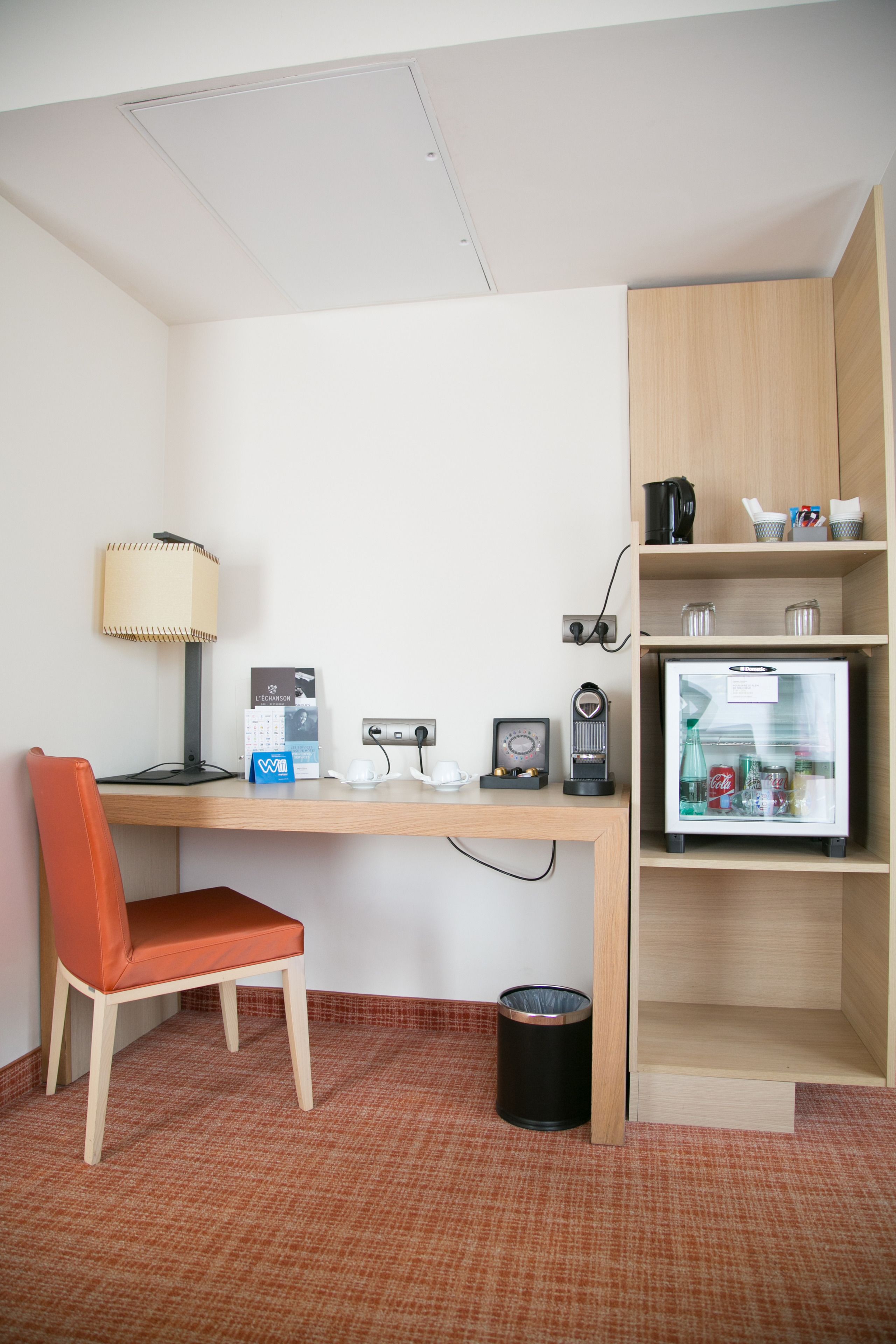 Una habitación de hotel compacta con escritorio, silla, lámpara, refrigerador y cesto de basura.