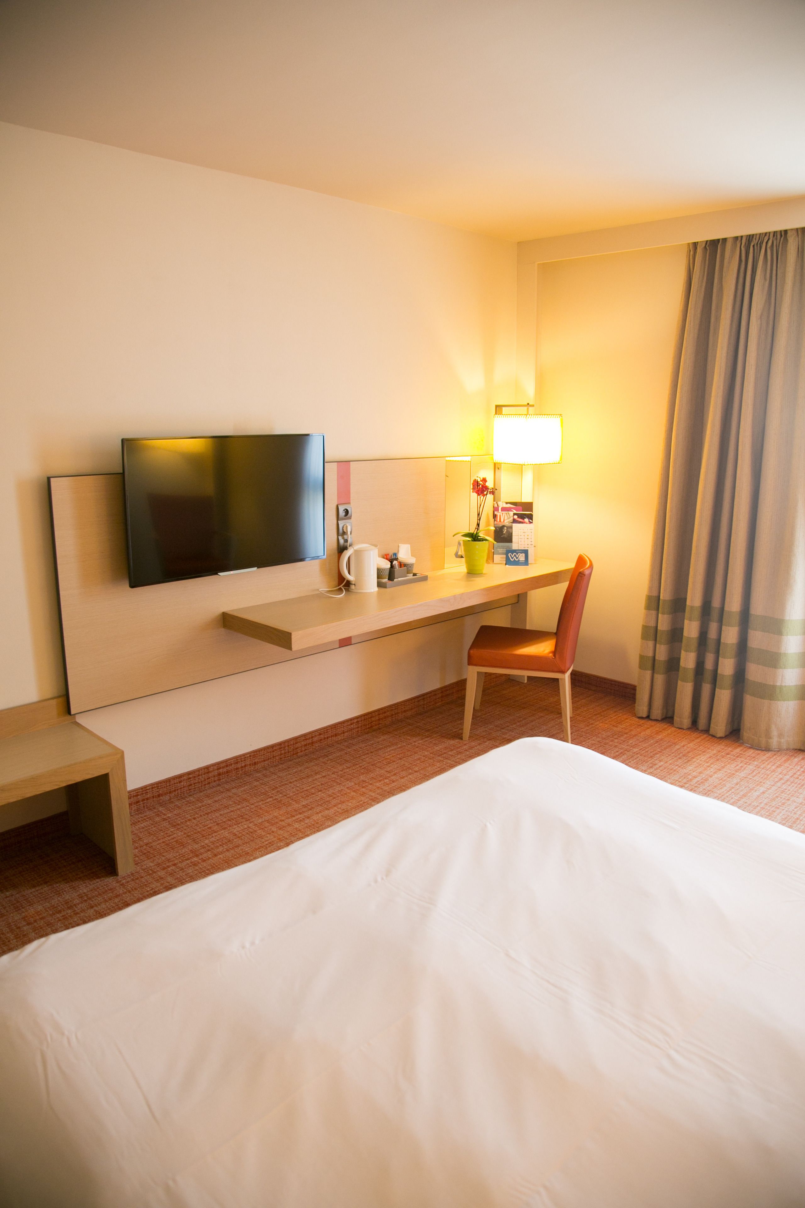 Habitación de hotel con cama, escritorio con silla, televisión, lámpara y ventana con cortinas.