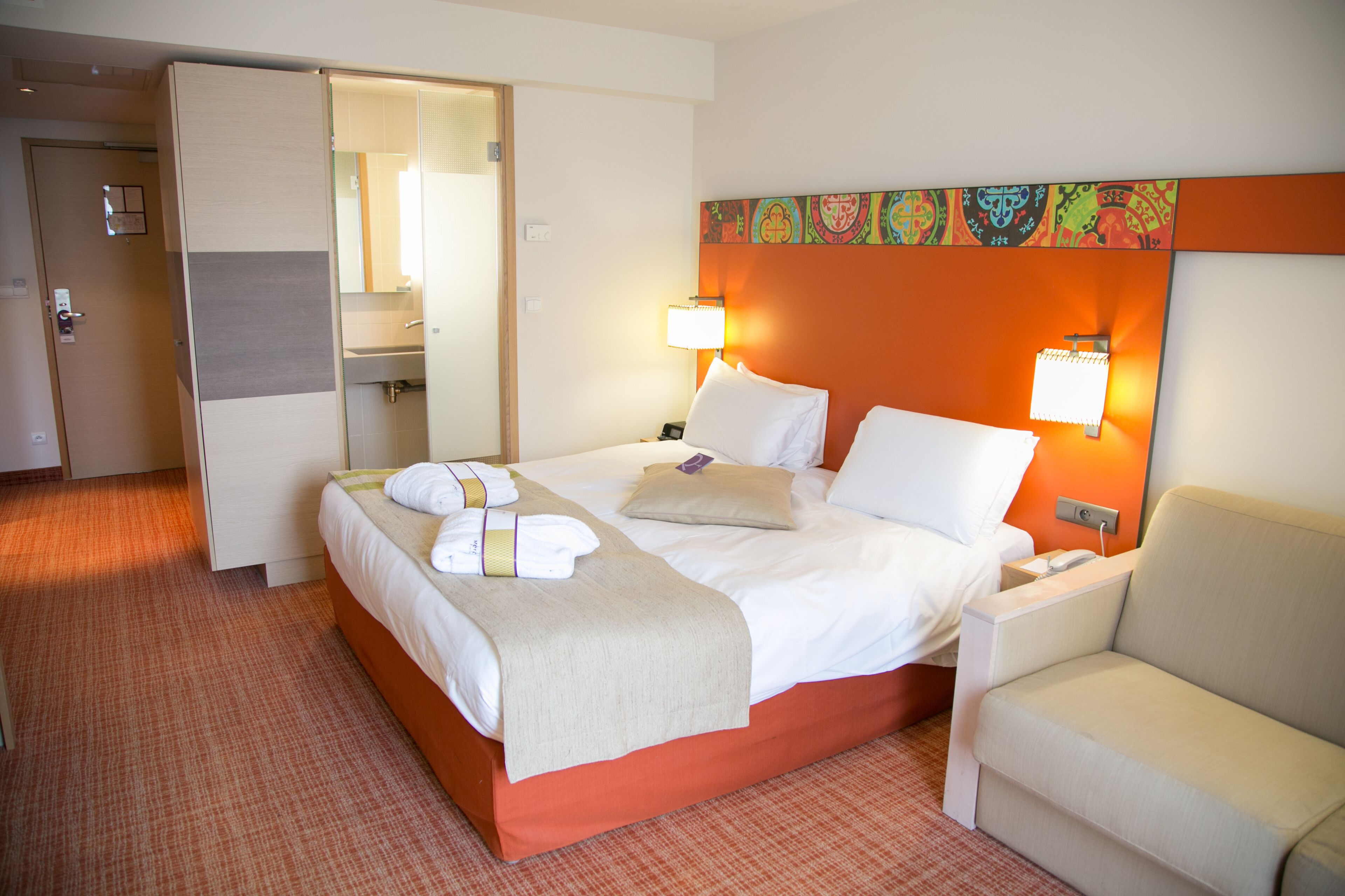 A hotel room with a bed, bedside tables, a chair, a bathroom, and a colorful wall decoration.