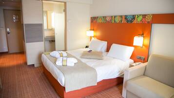 A hotel room with a bed, bedside tables, a chair, a bathroom, and a colorful wall decoration.