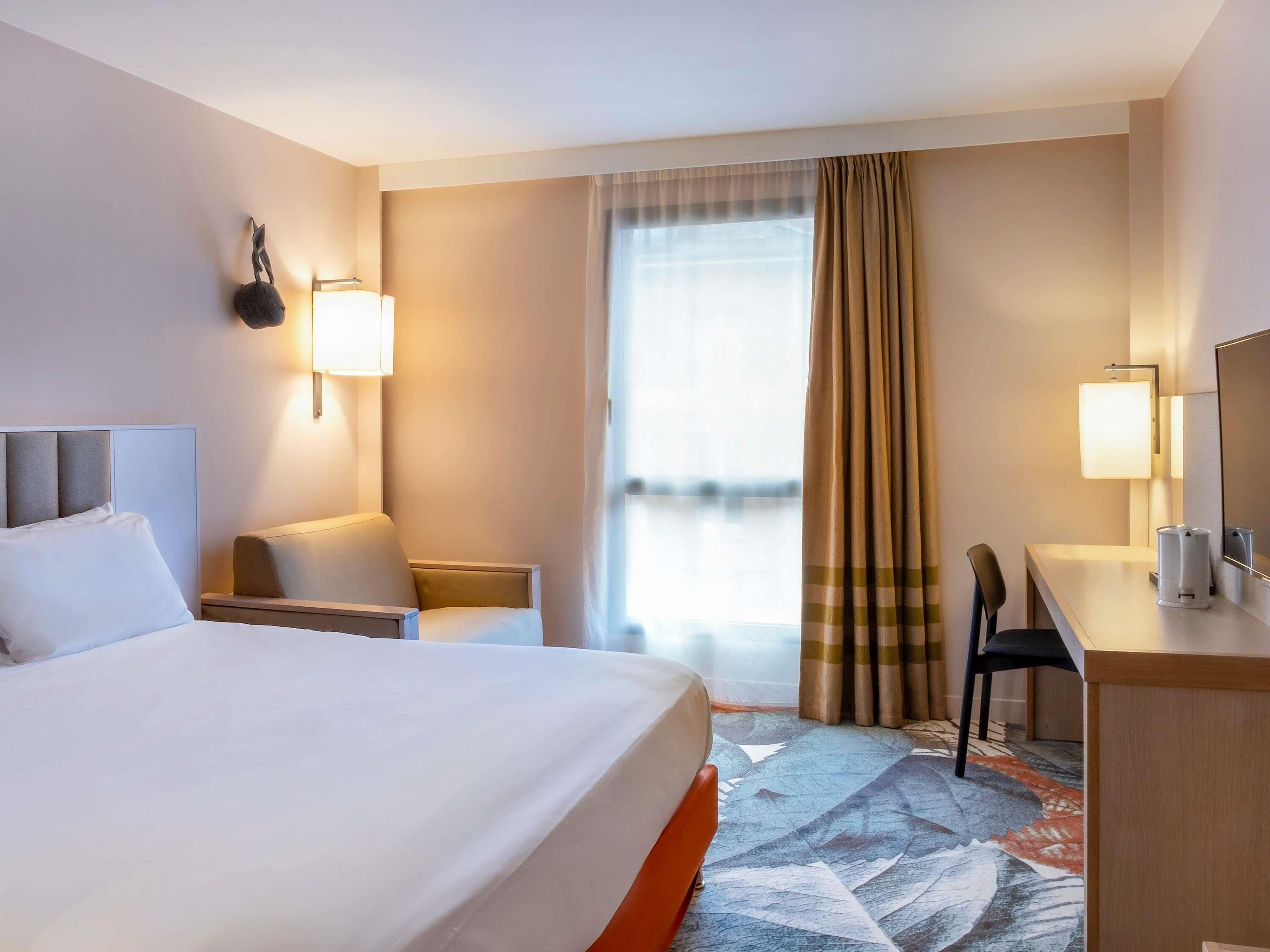 A hotel room with a large bed, a desk, a chair, a small table, a lamp, and a window with curtains.