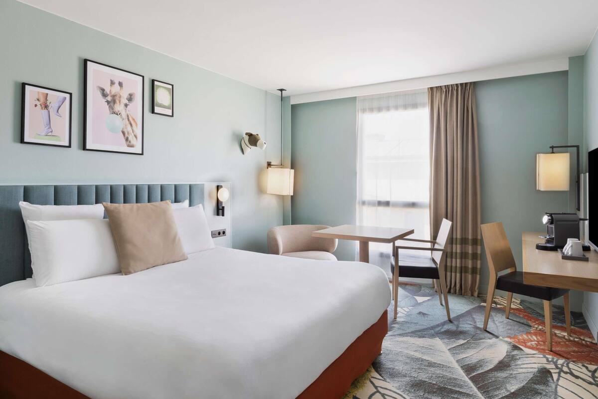 Ein Hotelzimmer mit einem großen Bett, einem Schreibtisch, einem Sessel, einem Fernseher und Bildern an den Wänden.