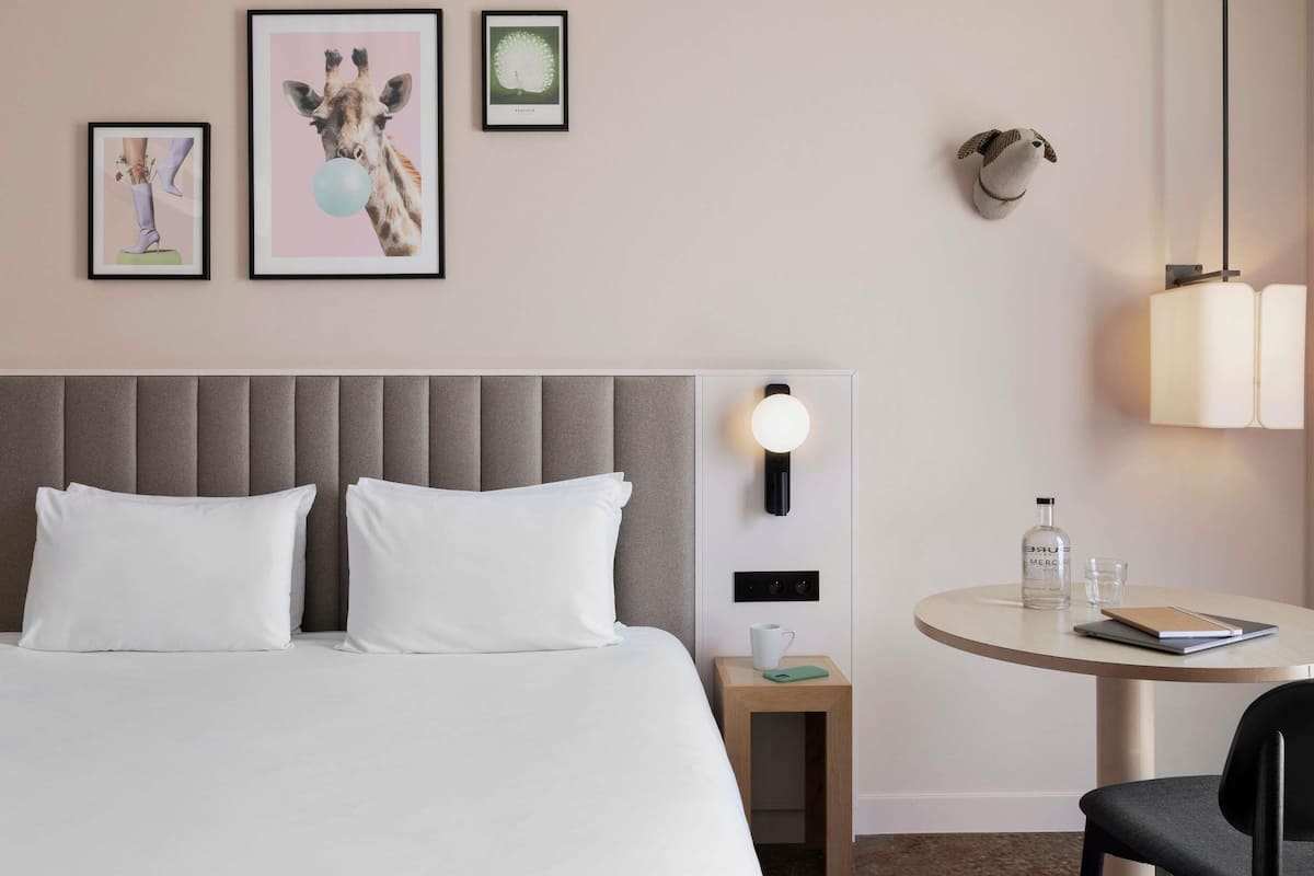 Ein Hotelzimmer mit Bett, Nachttisch, Tisch mit Stuhl und an der Wand befestigter Kunst.