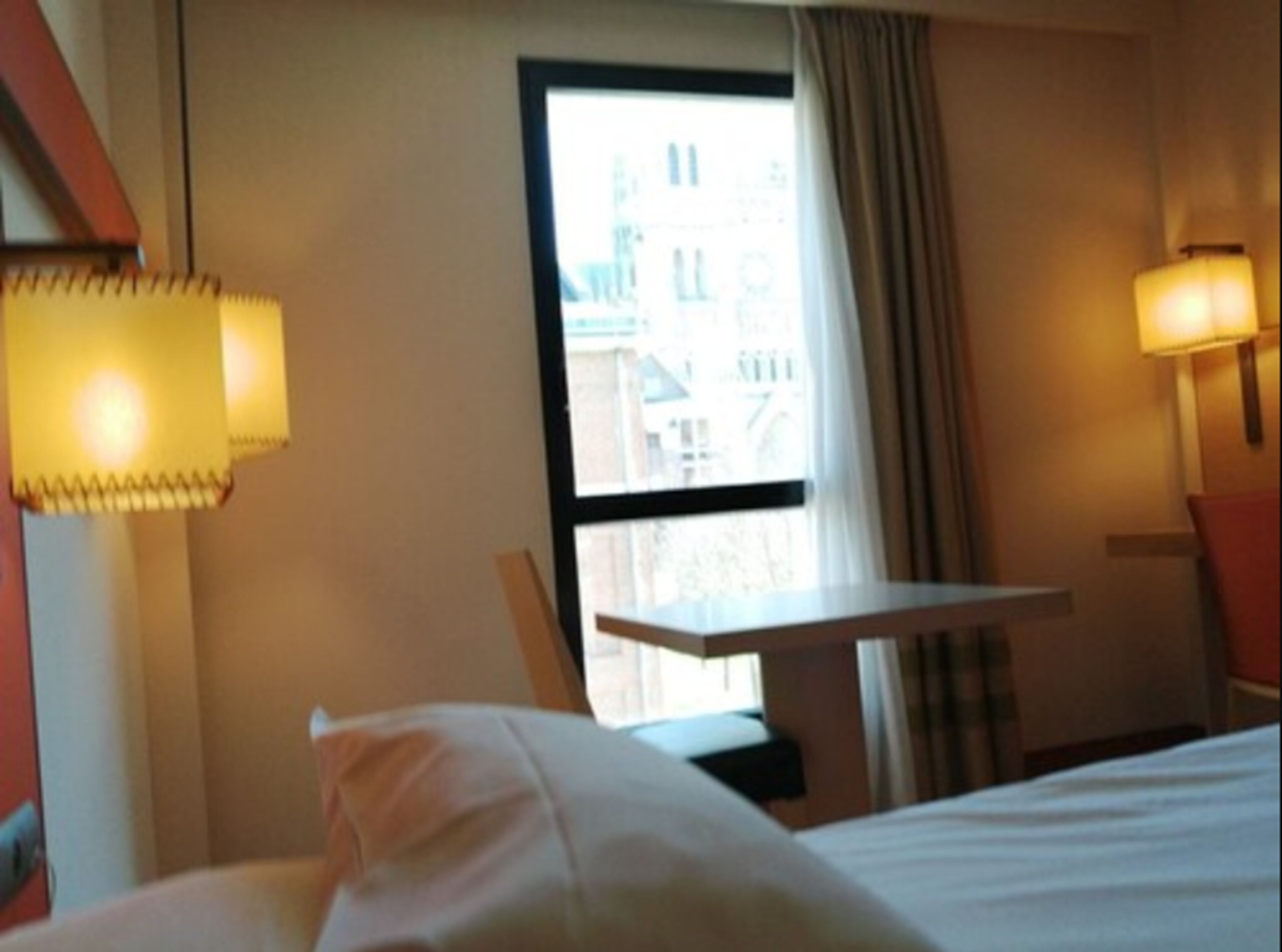 A hotel room with a bed, a small table, and a window with curtains.