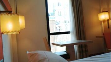 A hotel room with a bed, a small table, and a window with curtains.