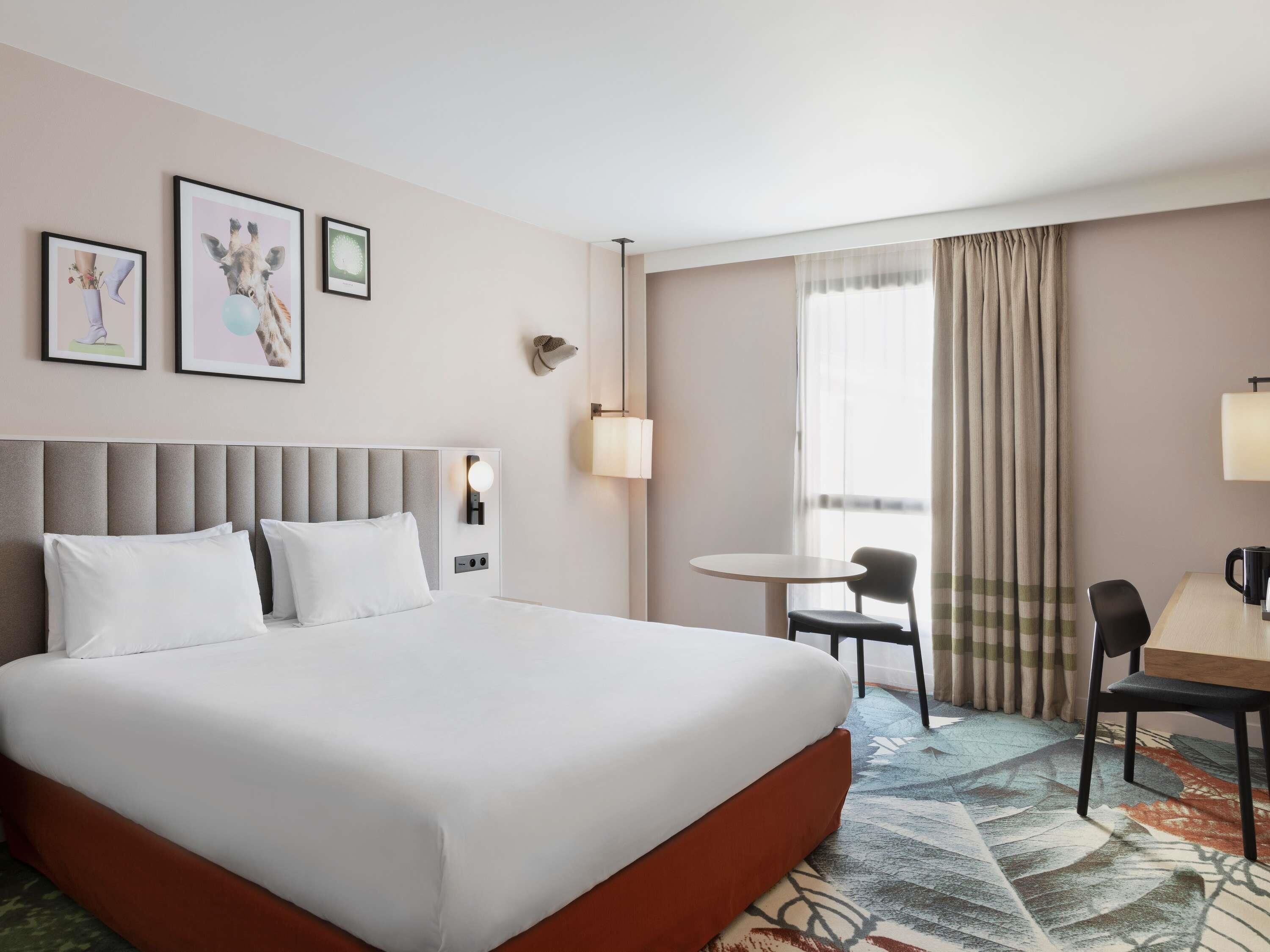 A hotel room with a large bed, a desk with a chair, a small table, and a window with curtains.