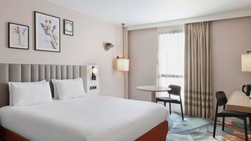 A hotel room with a large bed, a desk with a chair, a small table, and a window with curtains.