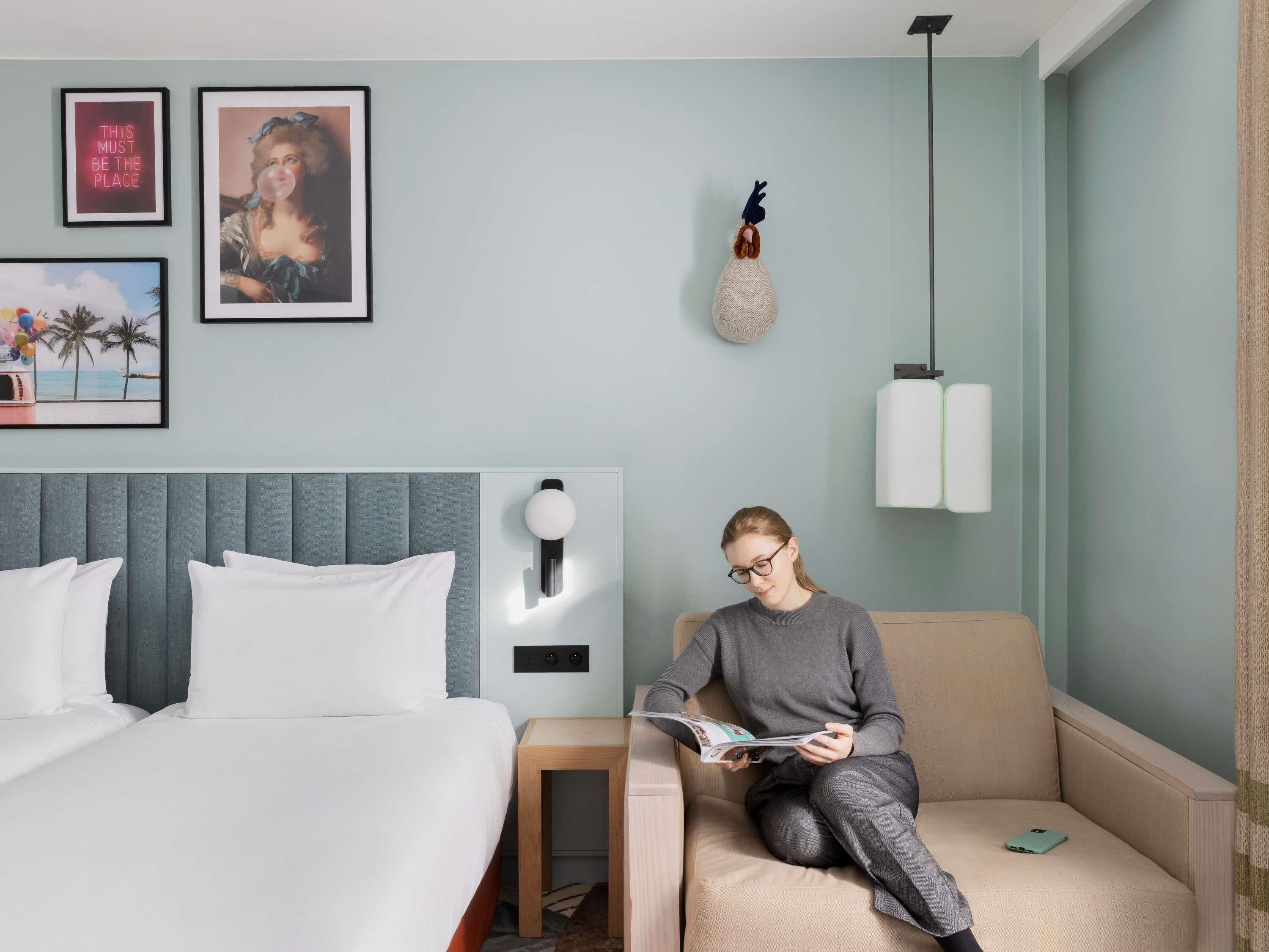 A hotel room with a bed, a reading chair, a small table, and wall-mounted light fixtures.
