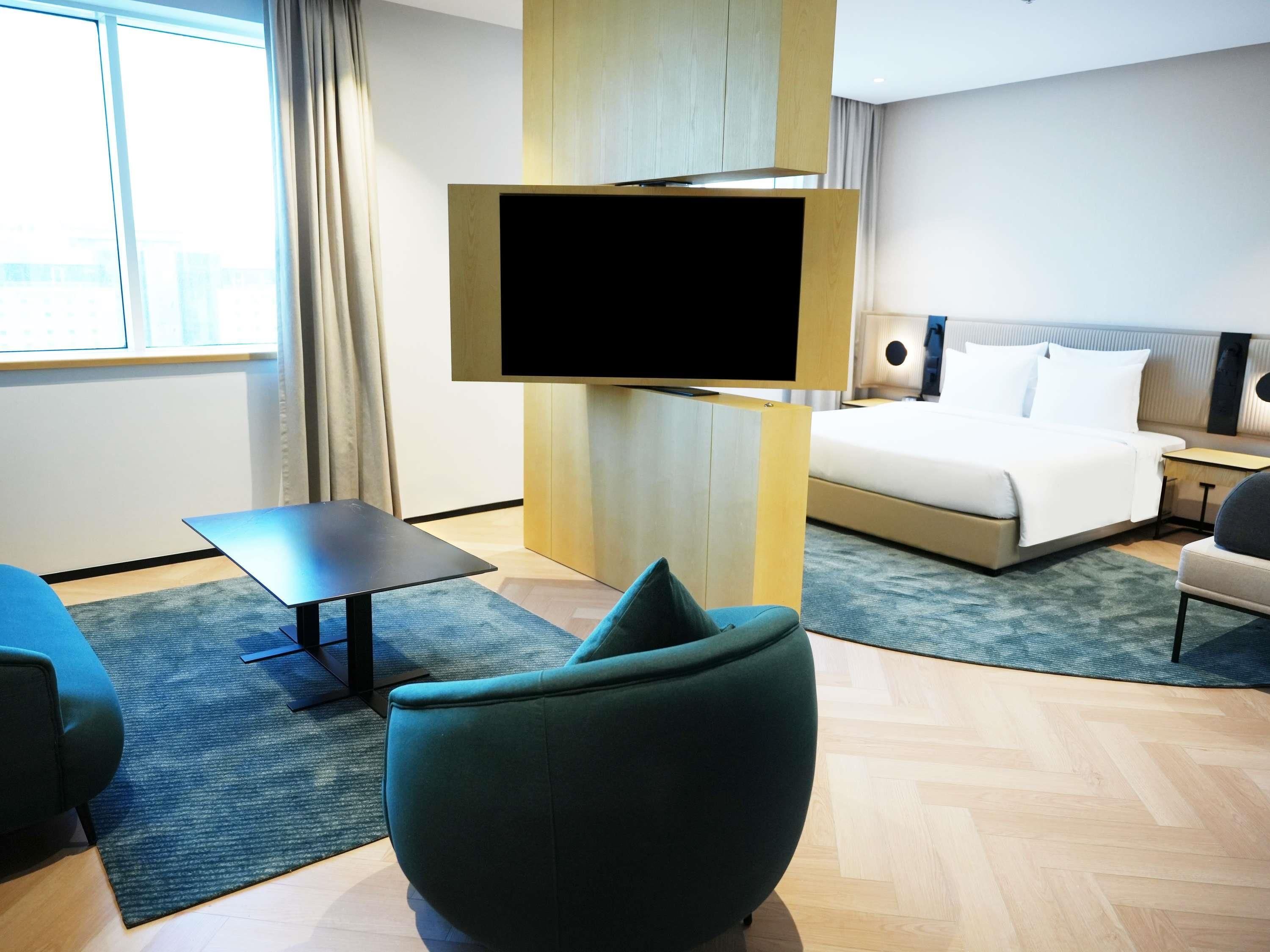 A modern hotel room with a large bed, a flat-screen TV mounted on a wooden panel, a small desk, and a comfortable armchair.