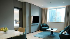 Een moderne hotelkamer met een flatscreen televisie aan de muur, een bank, een eettafel met stoelen, en uitzicht op de stad door grote ramen.