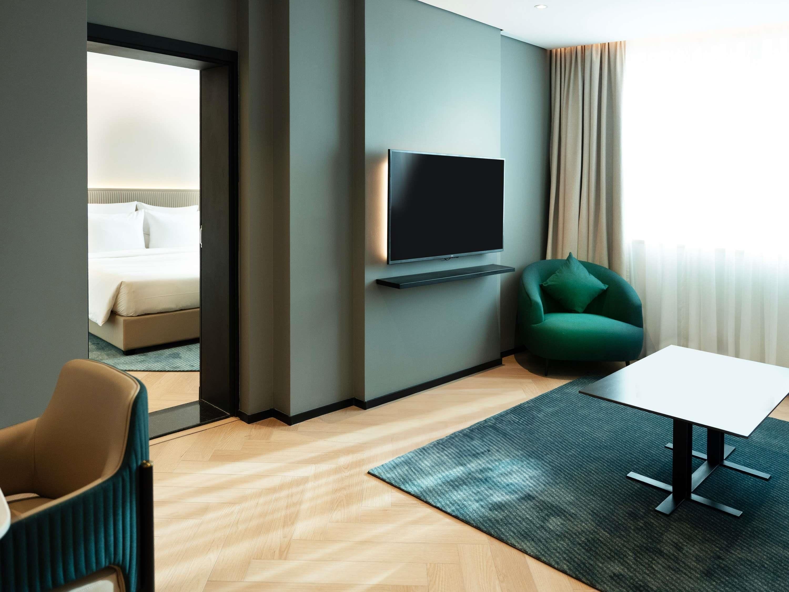 Une chambre d’hôtel moderne avec un lit, un téléviseur fixé au mur, un fauteuil vert, une petite table et une vue sur l’extérieur par une fenêtre avec des rideaux.