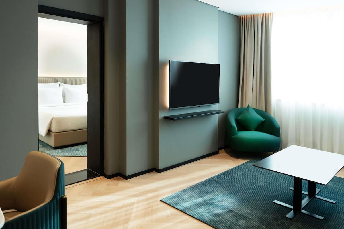 Une chambre d’hôtel moderne avec un lit, un téléviseur fixé au mur, un fauteuil vert, une petite table et une vue sur l’extérieur par une fenêtre avec des rideaux.